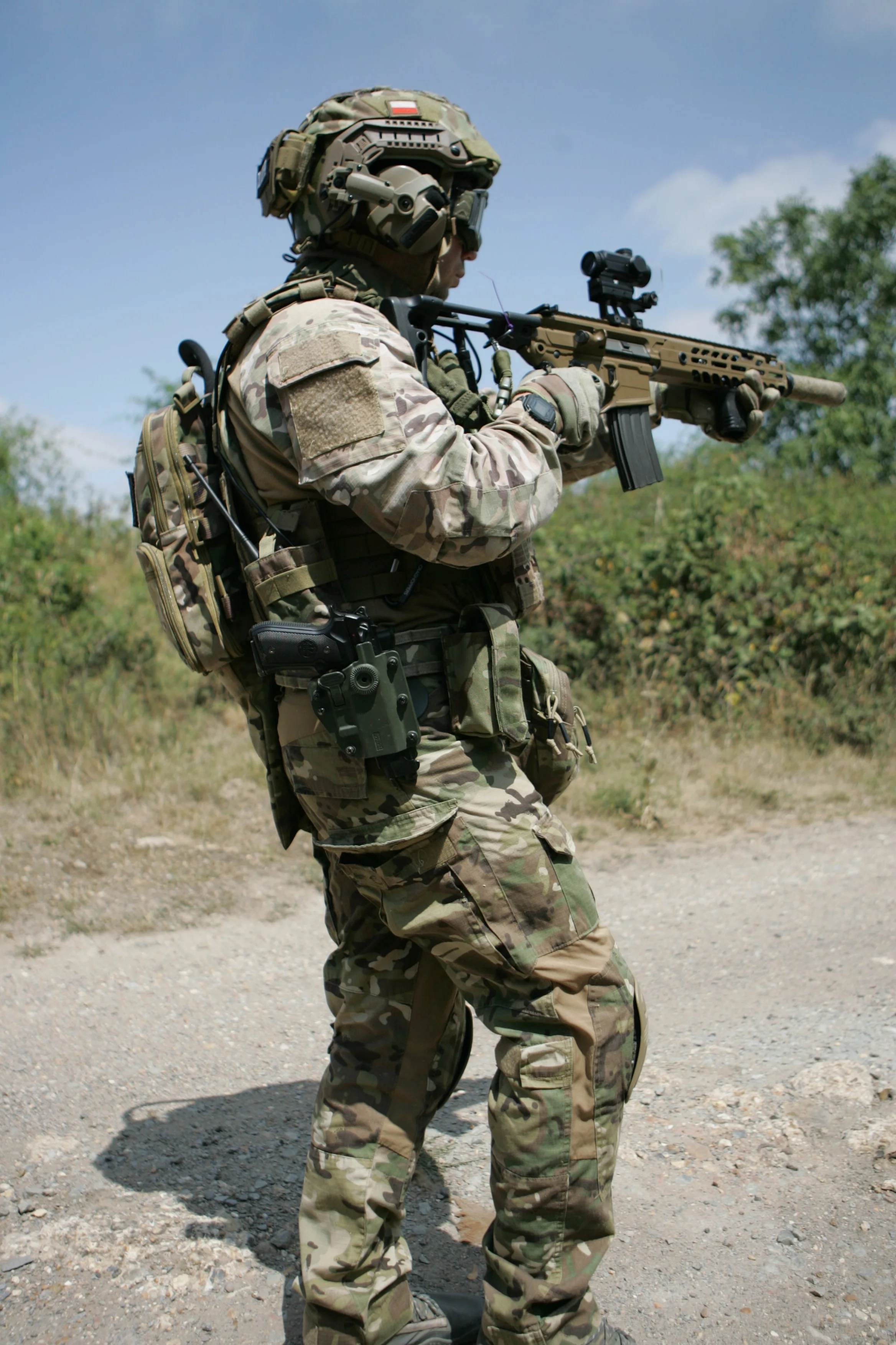 A soldier dressed in camouflage military uniform, wearing tactical gear, and aiming a rifle with a scope outdoors on a bright day with trees in the background.