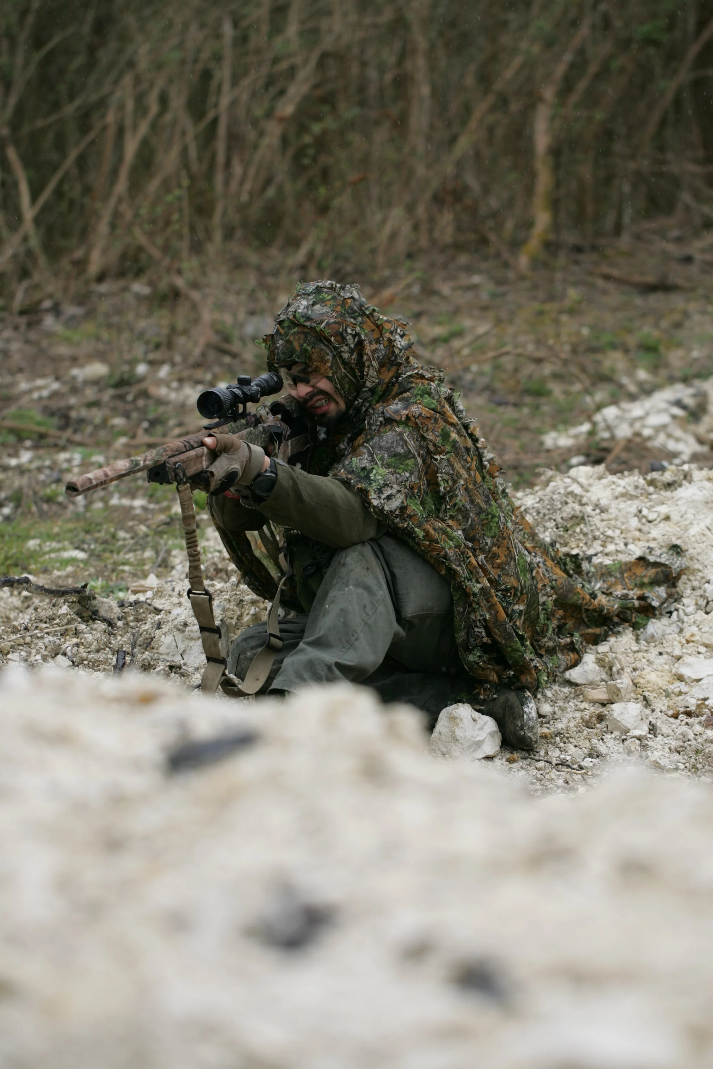 A person dressed in camouflage clothing and aiming a sniper rifle outdoors.