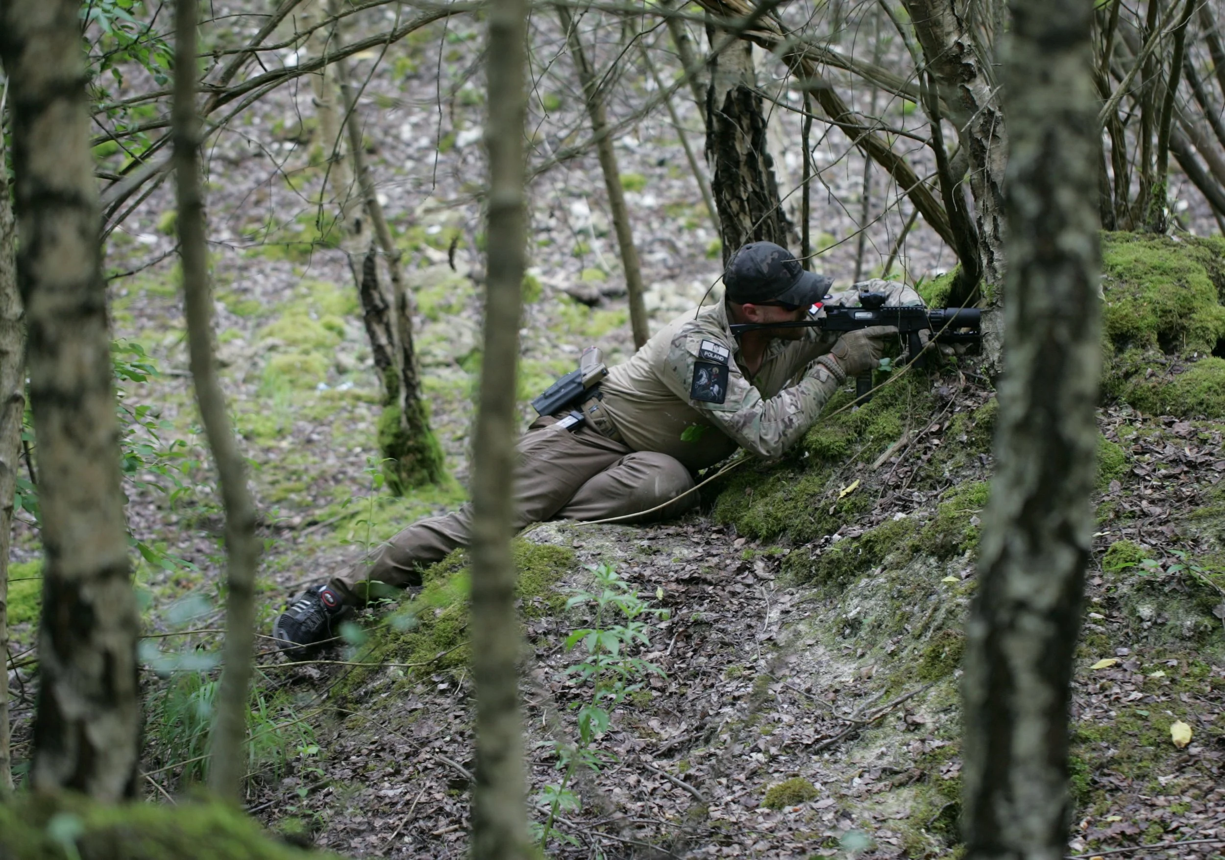 A soldier in camouflage uniform kneeling on the ground with a sniper rifle, aiming through a wooded forest.