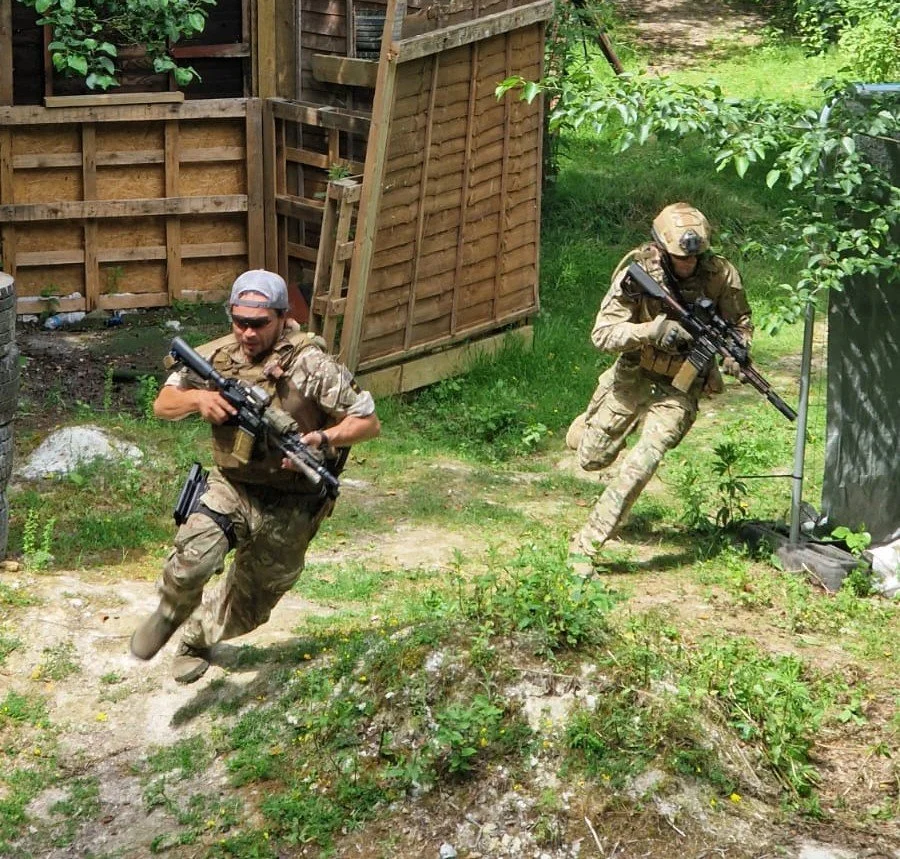 Two soldiers in camouflage gear running outdoors, one with a helmet and sunglasses, the other with a helmet and no sunglasses, both holding rifles.
