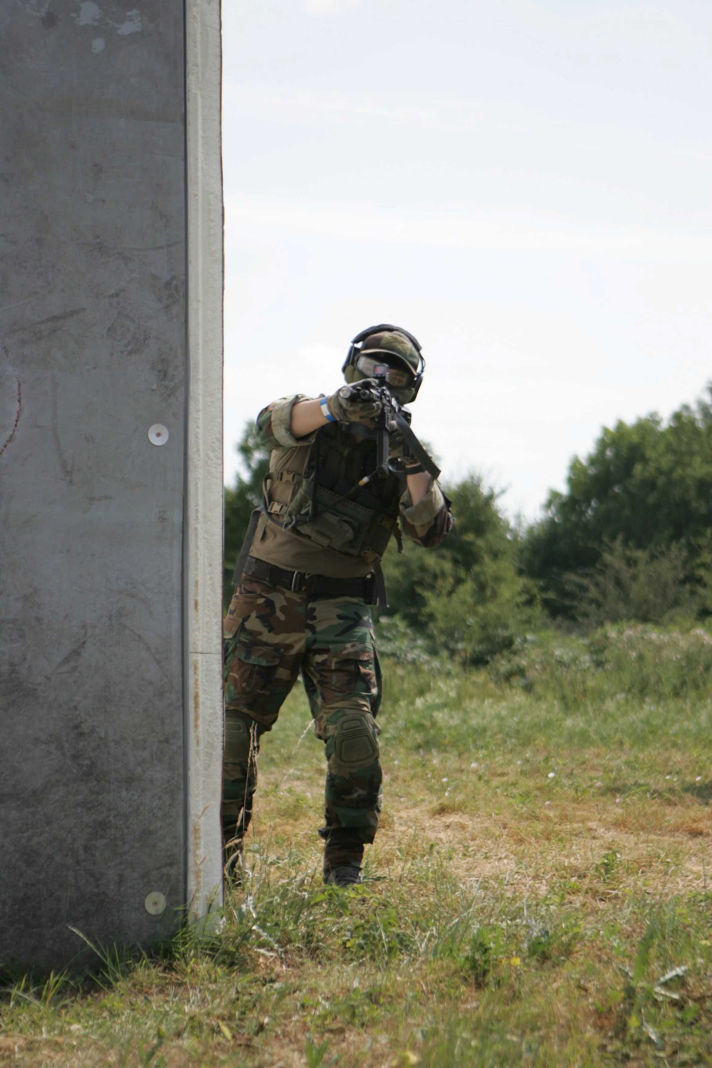 A person dressed in military camouflage and tactical gear crouches behind a concrete wall, aiming a firearm in an outdoor setting with trees and grass.