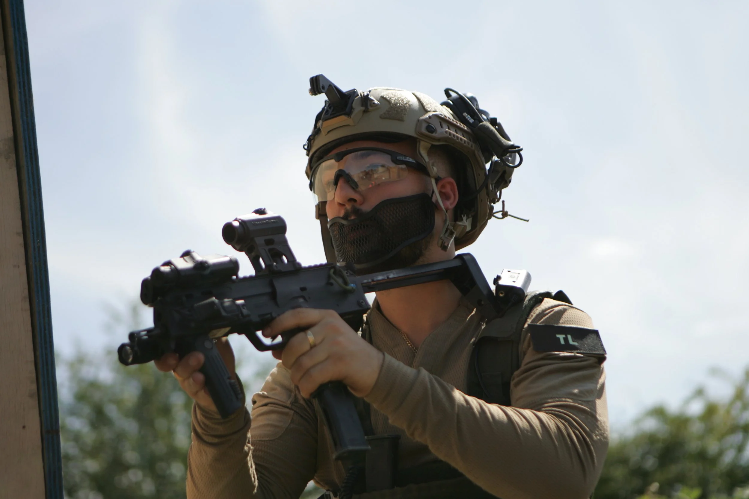 A soldier in tactical gear, including a helmet with mounted equipment, goggles, and a bulletproof vest, holding a handgun and aiming it during a training exercise or operation outdoors.