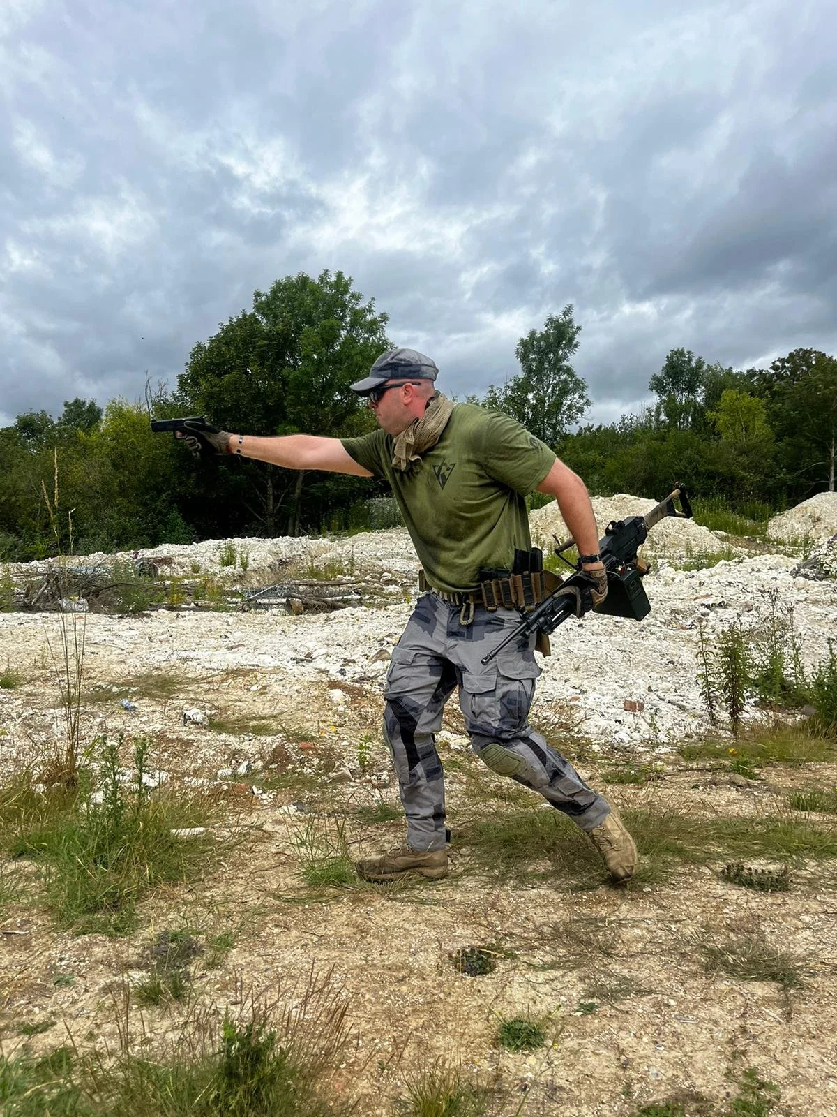 A man in tactical gear holding a handgun in a shooting stance outdoors on a cloudy day, with trees and open land in the background.