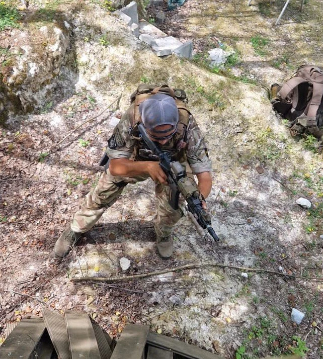Soldier in camouflage uniform and cap crouching while aiming a rifle in a wooded area surrounded by dirt and rocks.