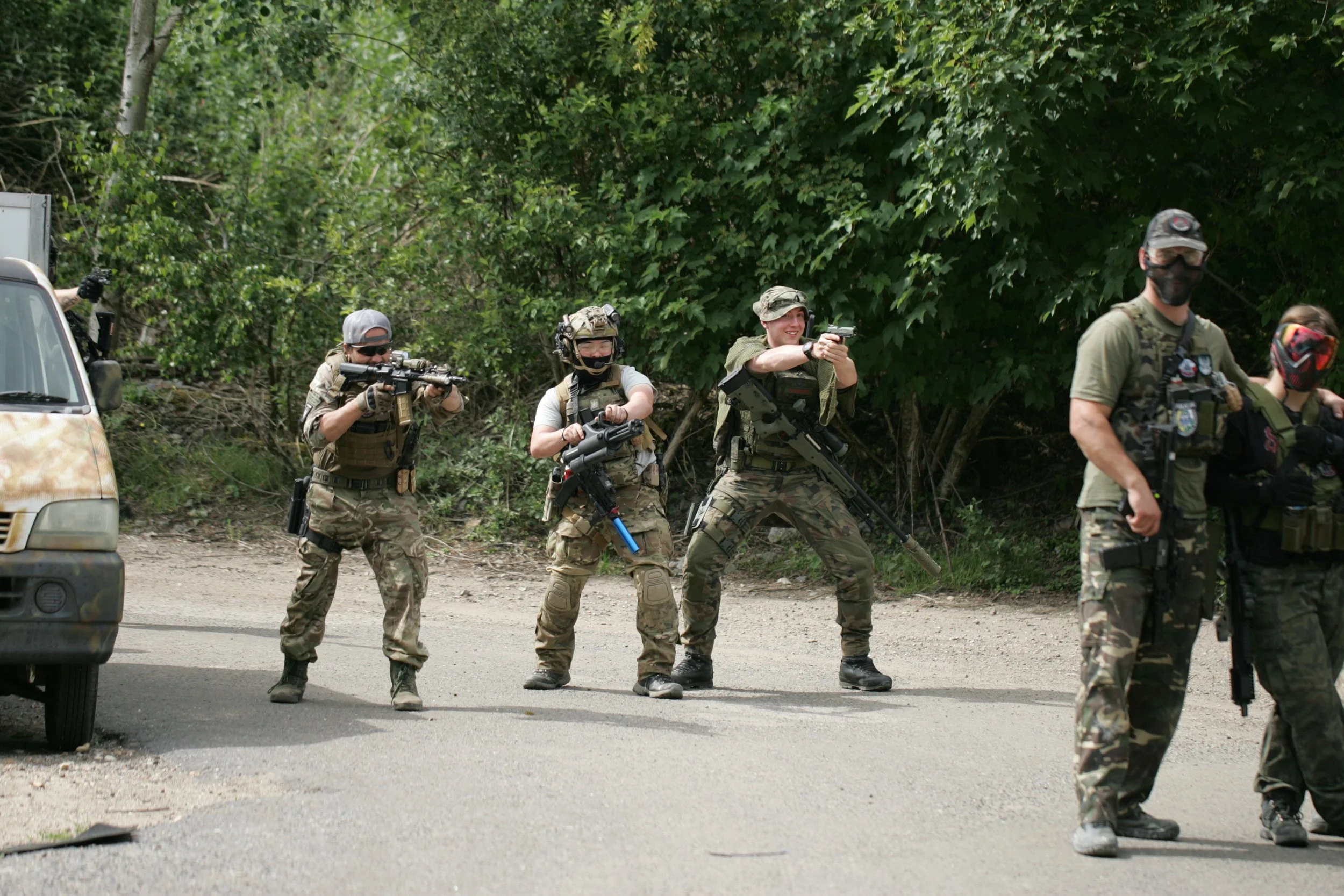Group of people in military gear practicing firearm training outdoors with trees and greenery in the background.