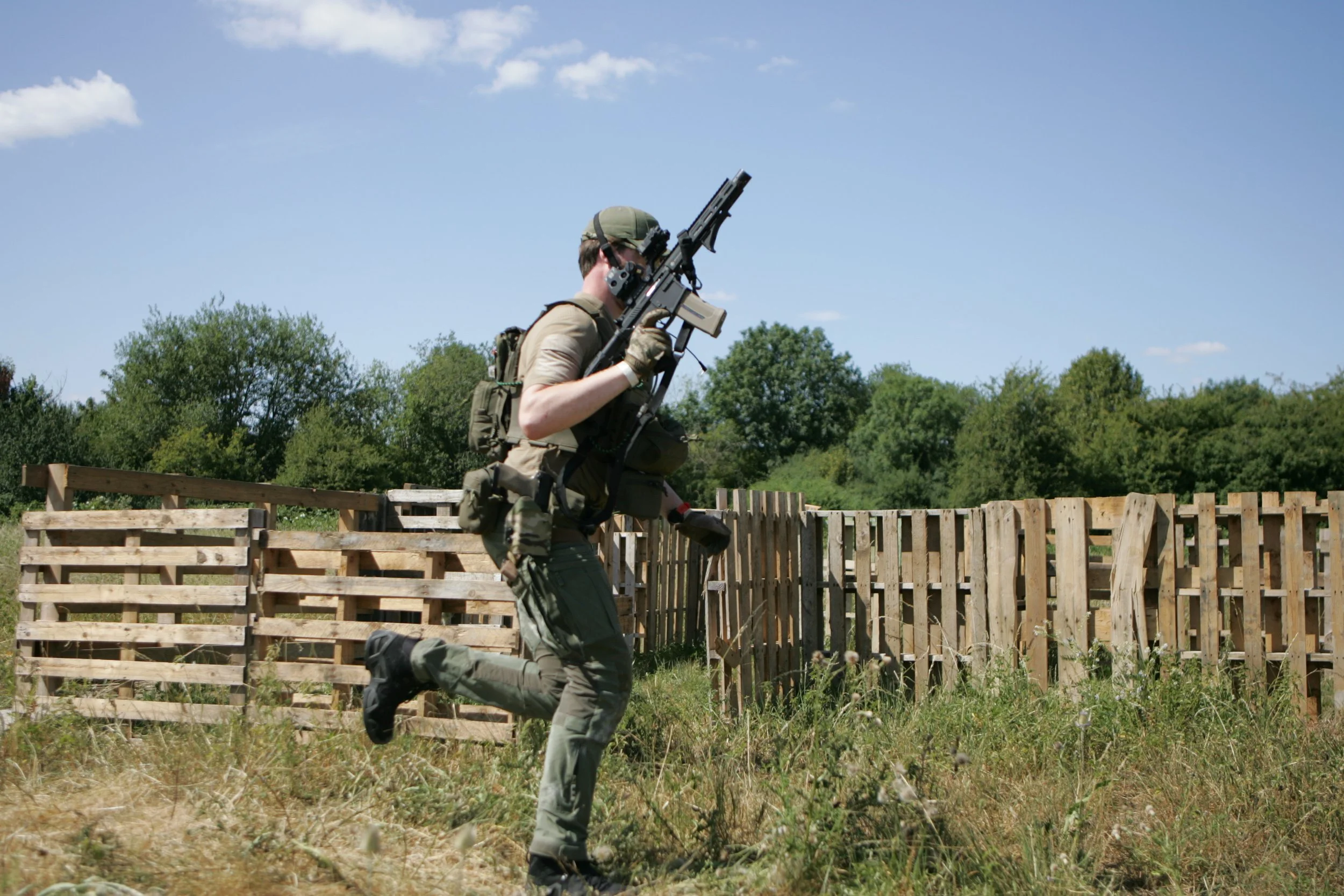 A soldier running outdoors next to a wooden fence, holding a rifle, with trees and a blue sky in the background.