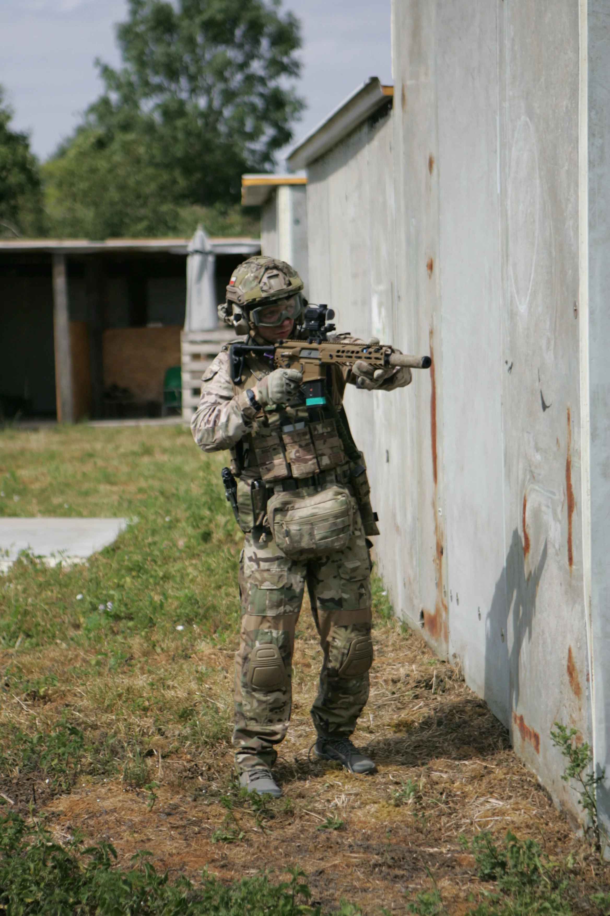 A soldier in camouflage uniform and tactical gear aiming a rifle behind a concrete wall on a shooting range or training ground.