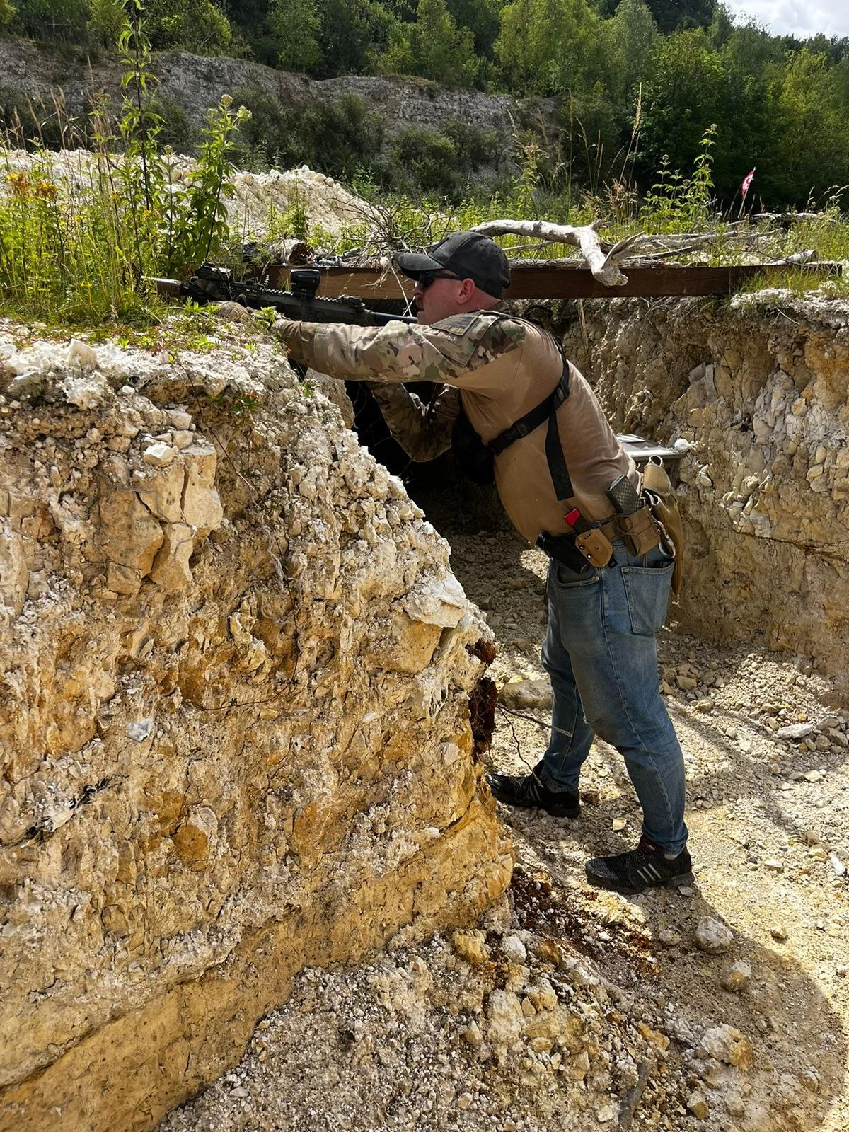 A man in camouflage and jeans, wearing a black cap and sunglasses, aiming a scoped rifle through a narrow excavation in rocky terrain with trees in the background.