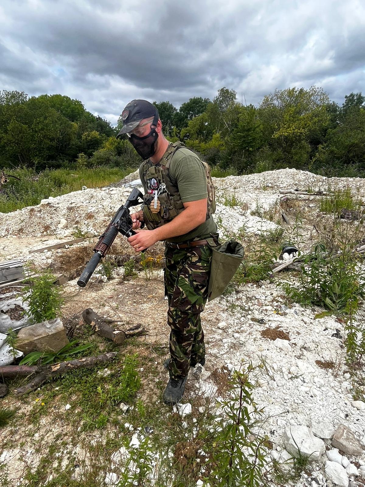 A soldier in camouflage pants, a green t-shirt, and tactical gear, standing outdoors on rocky terrain with greenery and trees in the background. The soldier is holding a rifle and wearing a face mask and cap, with a cloudy sky overhead.