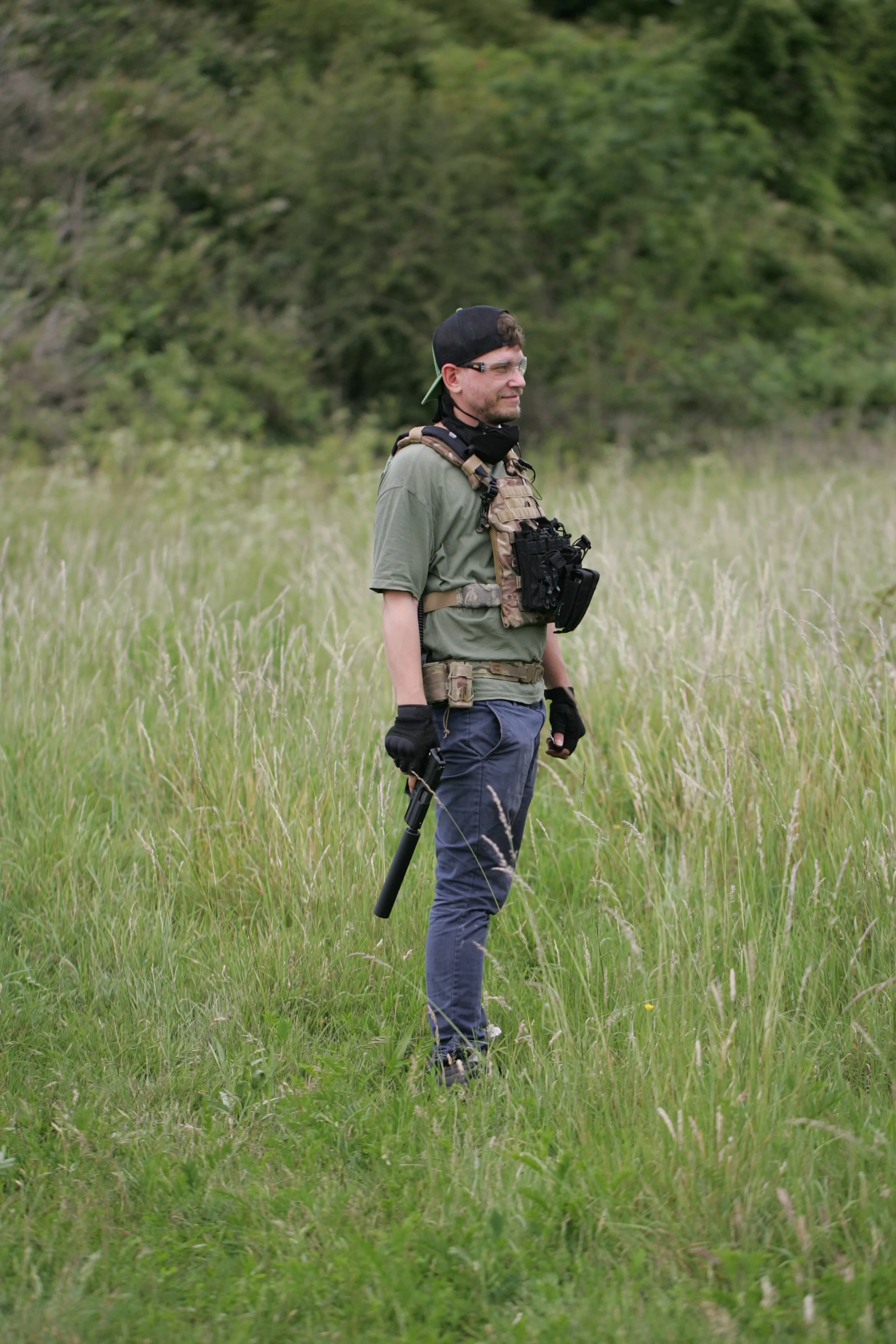 A man wearing glasses, a backward cap, tactical gear, and gloves stands in a grassy field with trees in the background, holding a metal detector.