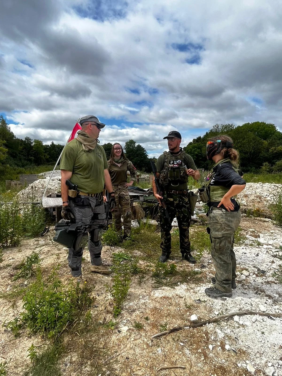 Four individuals dressed in tactical gear, standing outdoors on rocky terrain, engaged in conversation with lush green trees and cloudy sky in the background.