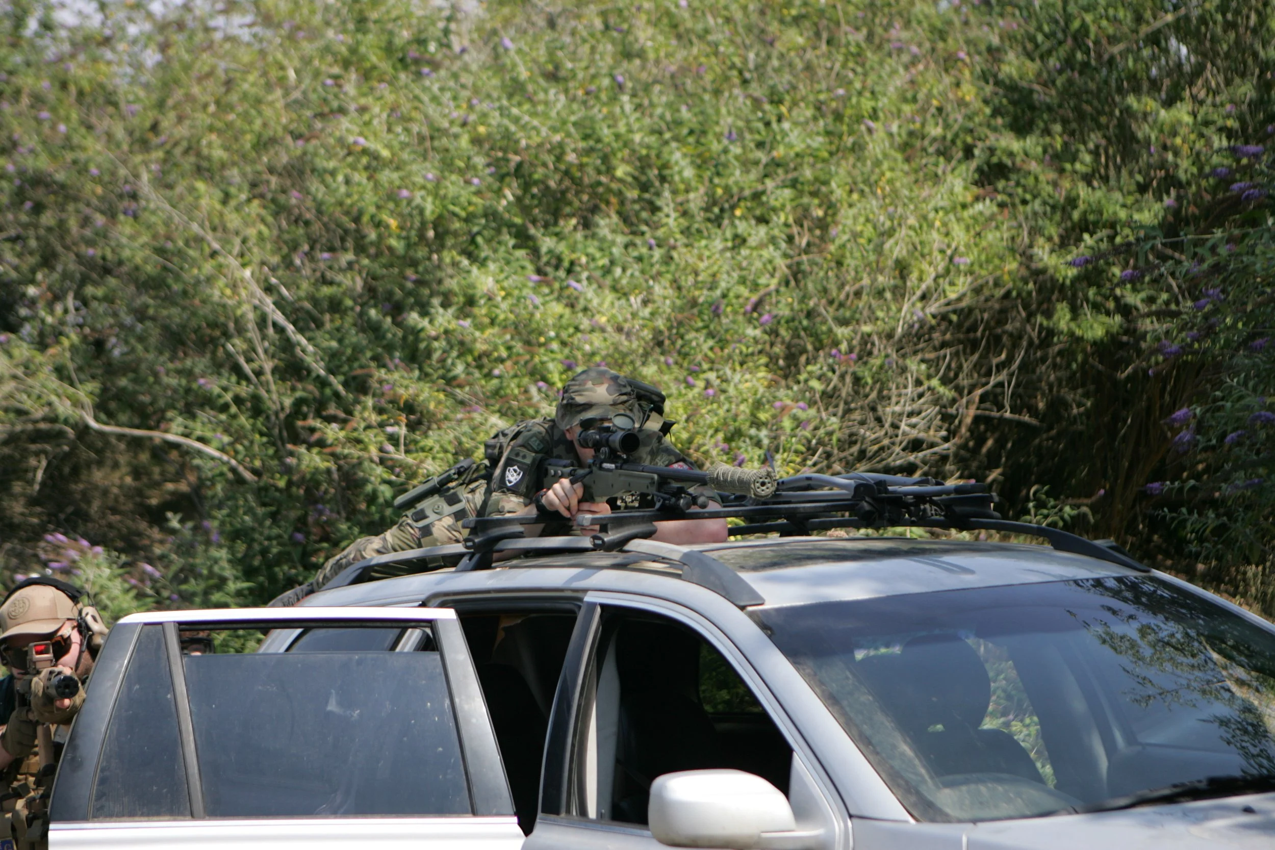A soldier in camouflage gear aiming a sniper rifle from the roof of a vehicle amidst dense bushes.