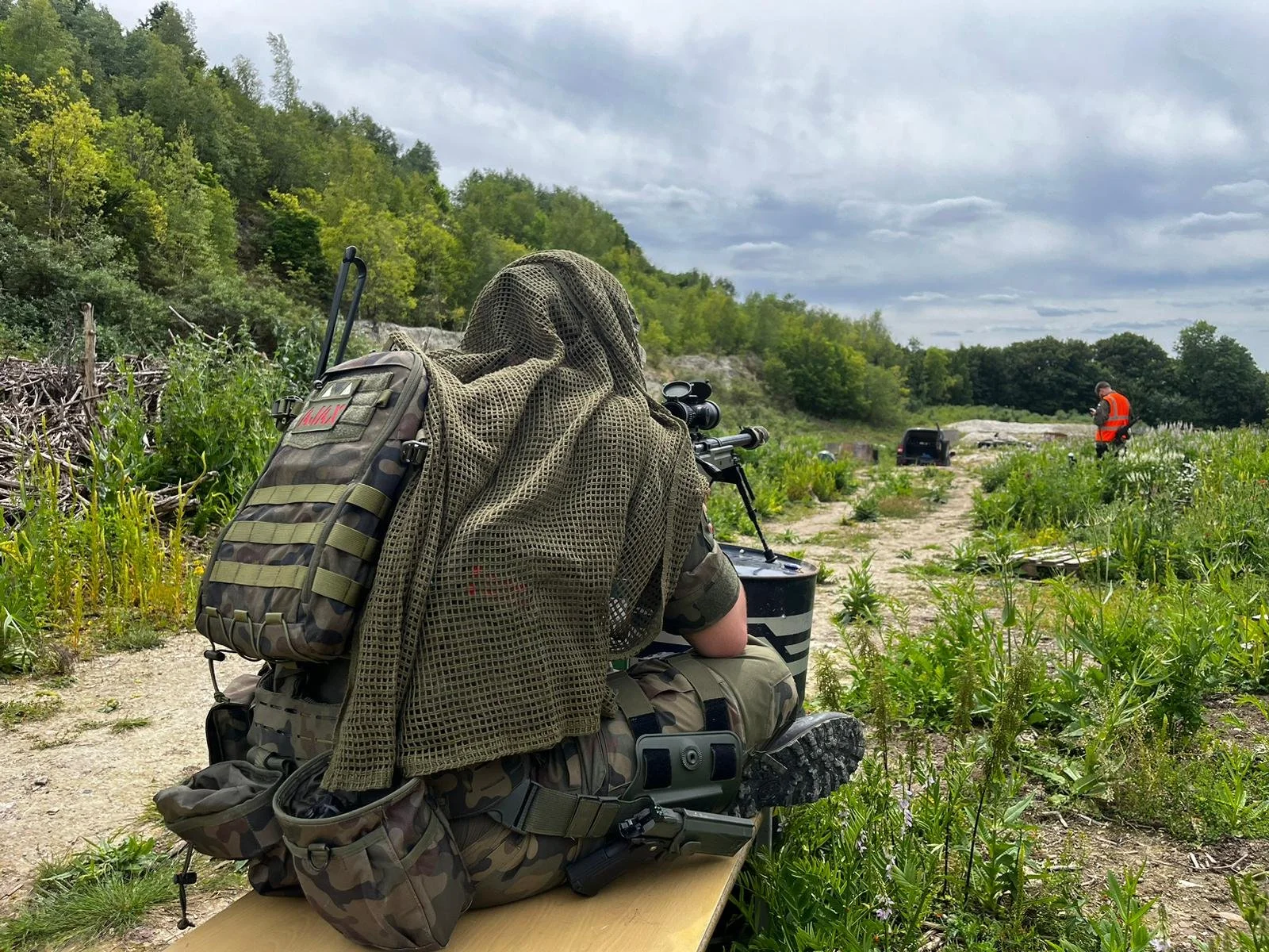 Person in camouflage clothing and tactical gear sitting on a scouting vehicle with a scoped rifle, observing an outdoor wooded area with another person in an orange vest in the distance.