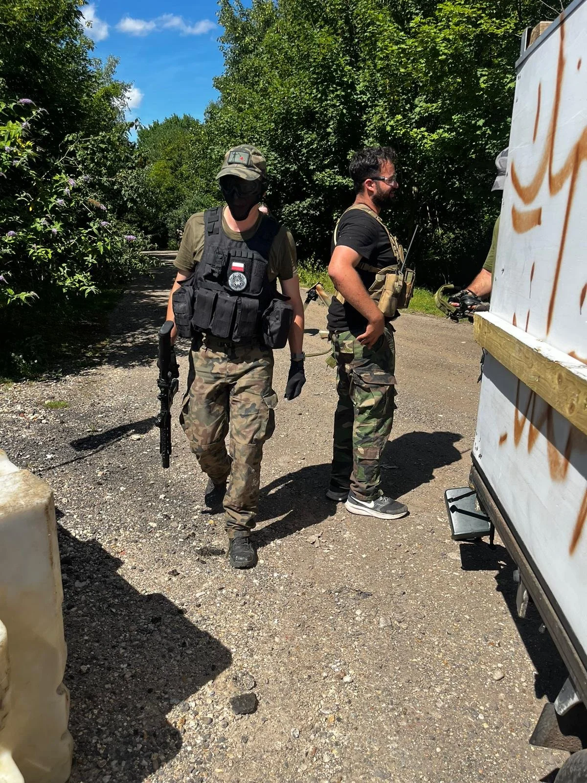 Two individuals dressed in camouflage military attire near a white trailer, with one holding a rifle, in a wooded outdoor setting.