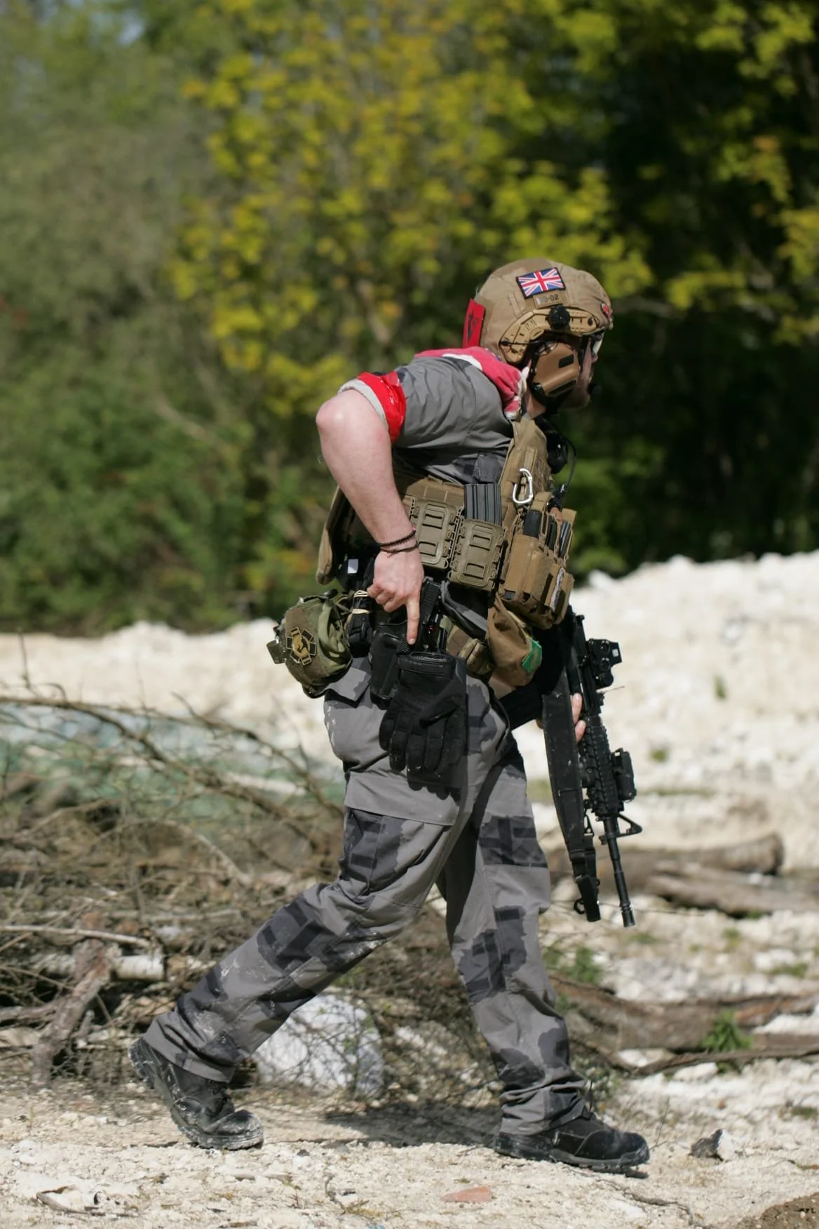 A soldier in tactical gear walking outdoors on a dirt surface, with a green forest in the background, holding a rifle.
