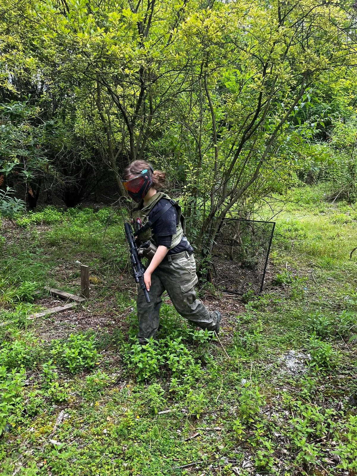 A person in camouflage pants, a black shirt, and protective gear including a helmet and goggles, holding a firearm, and standing in a green, wooded outdoor area.