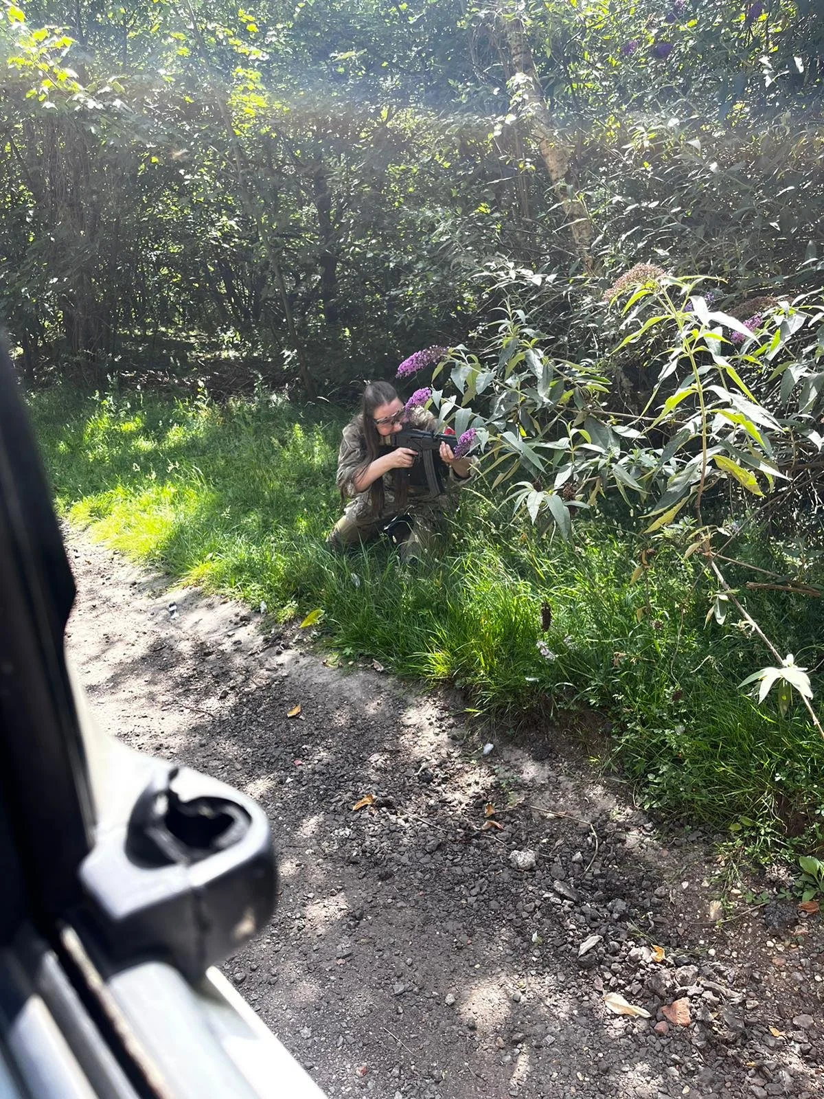 A woman in military camouflage gear crouches in a wooded area, aiming a rifle through bushes with purple flowers.