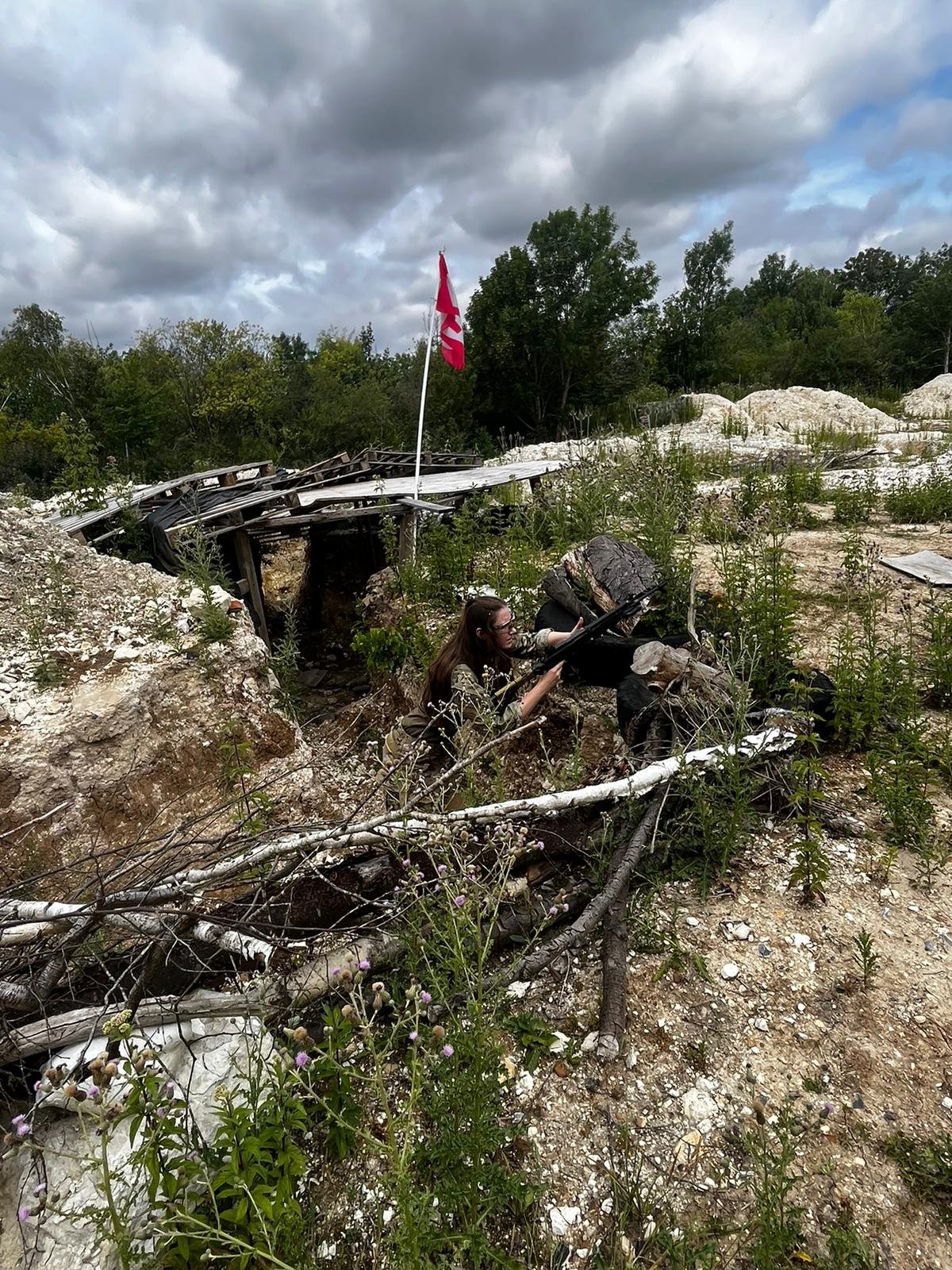 A woman in military camouflage takes cover behind rocks and bushes with a rifle, in a damaged outdoor area with debris, greenery, and a cloudy sky.