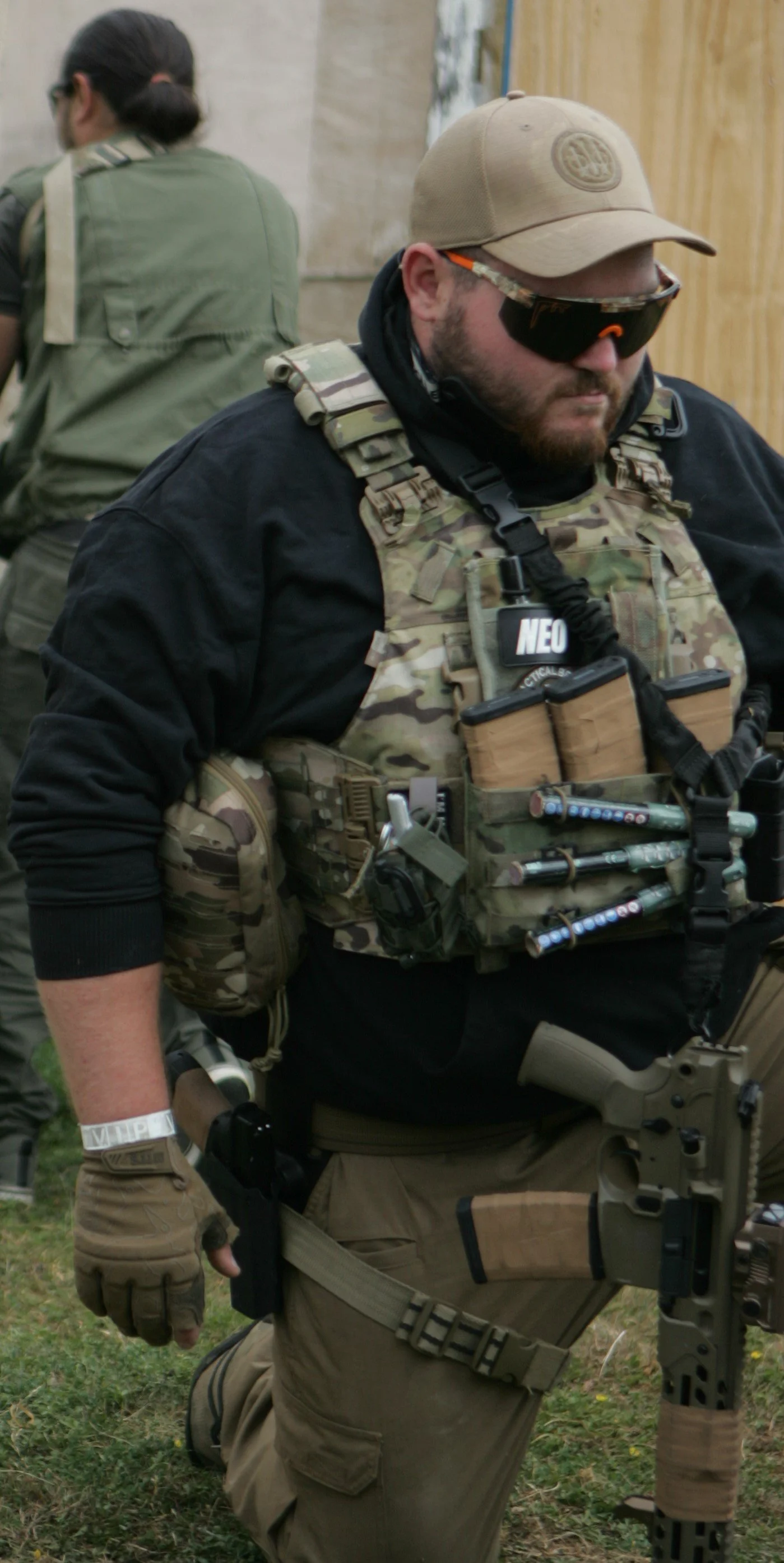 Man dressed in tactical gear with a tactical vest, goggles, and a cap, kneeling outdoors with a firearm.