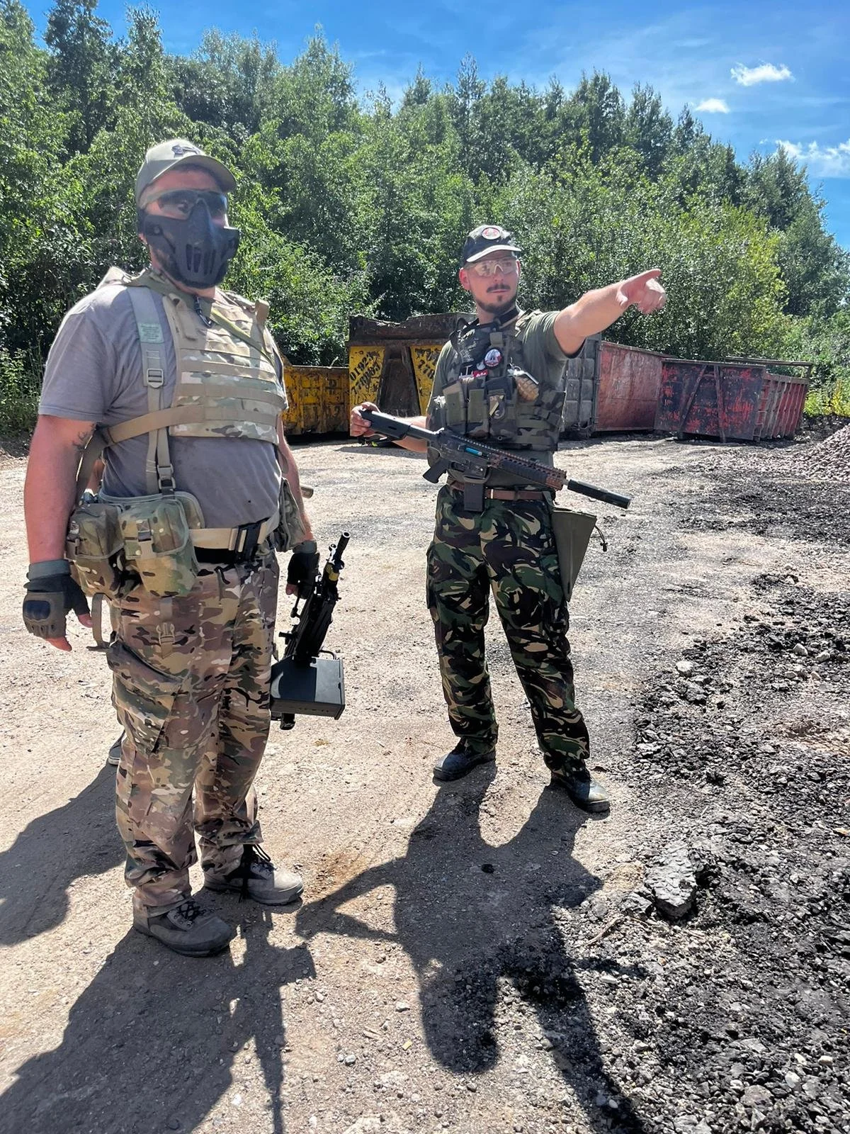 Two men in military camouflage uniforms with tactical gear and weapons standing outdoors on a dirt surface, with trees and rusted dumpsters in the background. One man is pointing.