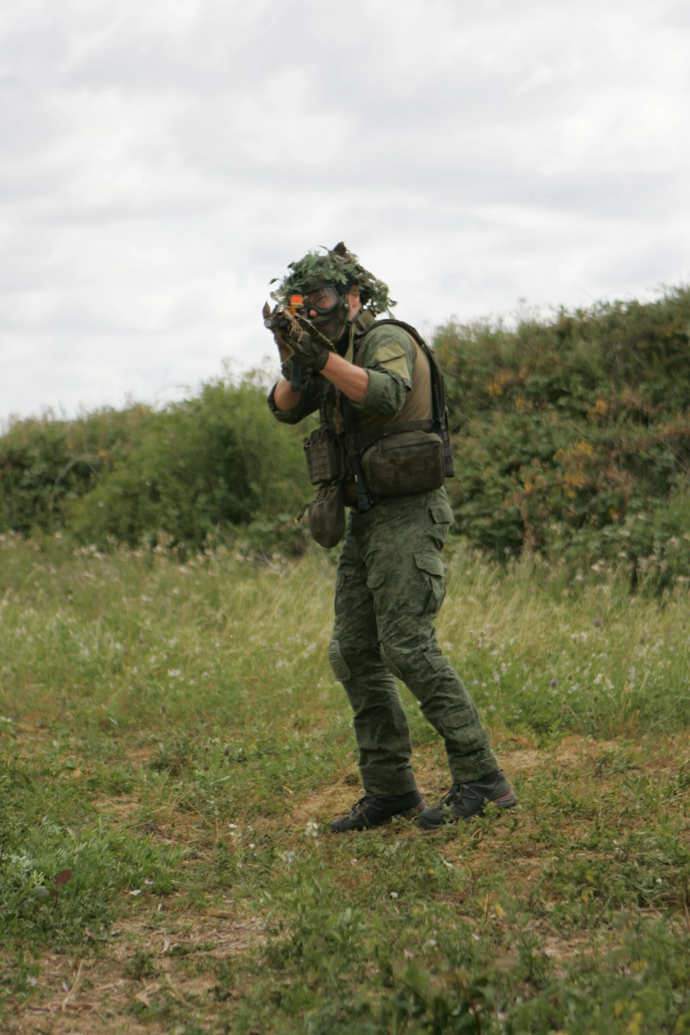 Soldier in camouflage uniform and tactical gear crouches on grassy terrain, aiming a rifle with shrubbery and cloudy sky in the background.