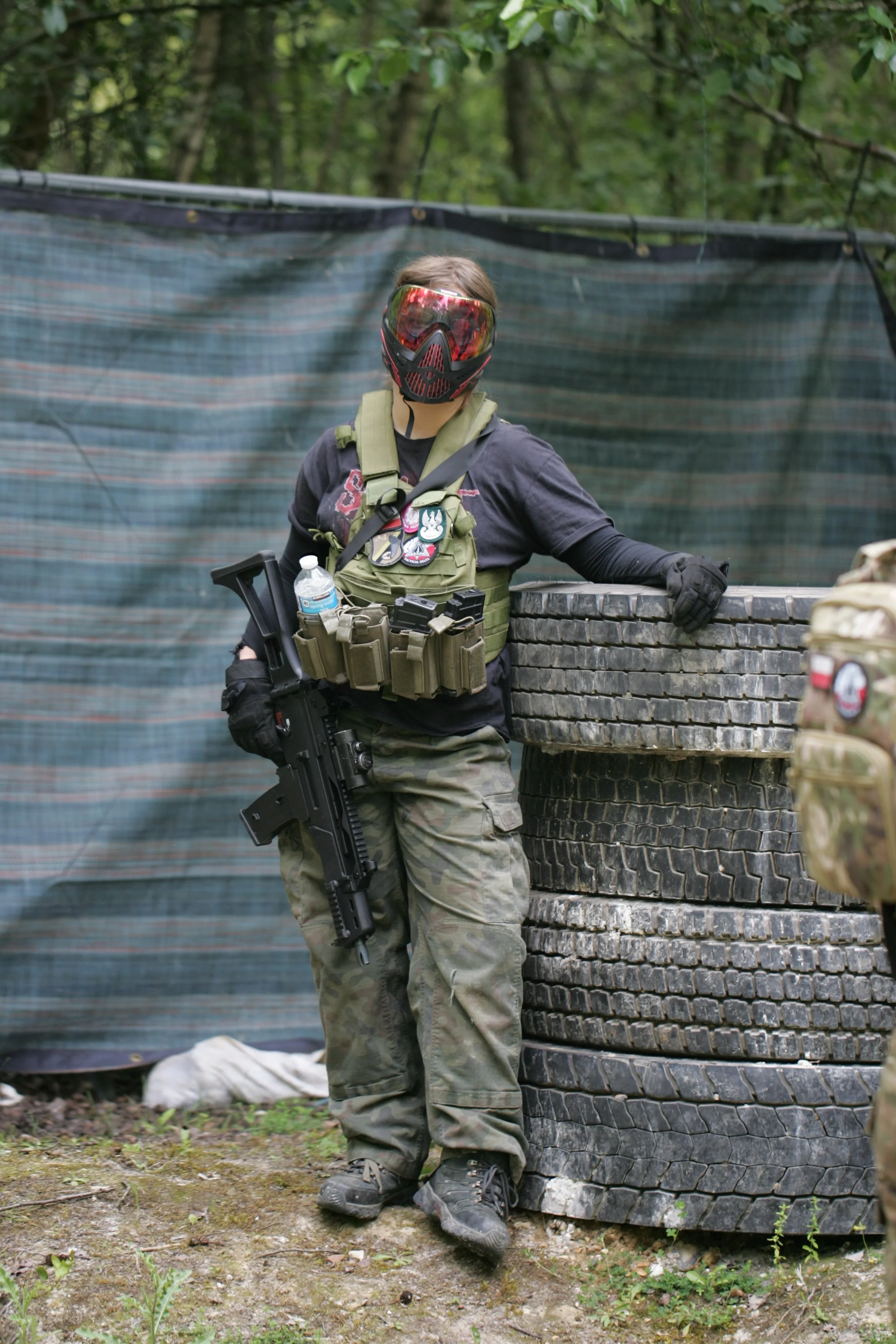 Person wearing tactical gear, including camouflage pants, black gloves, and a vest with patches, standing next to a stack of tires and holding a firearm in a wooded area with a black and blue striped barrier in the background.