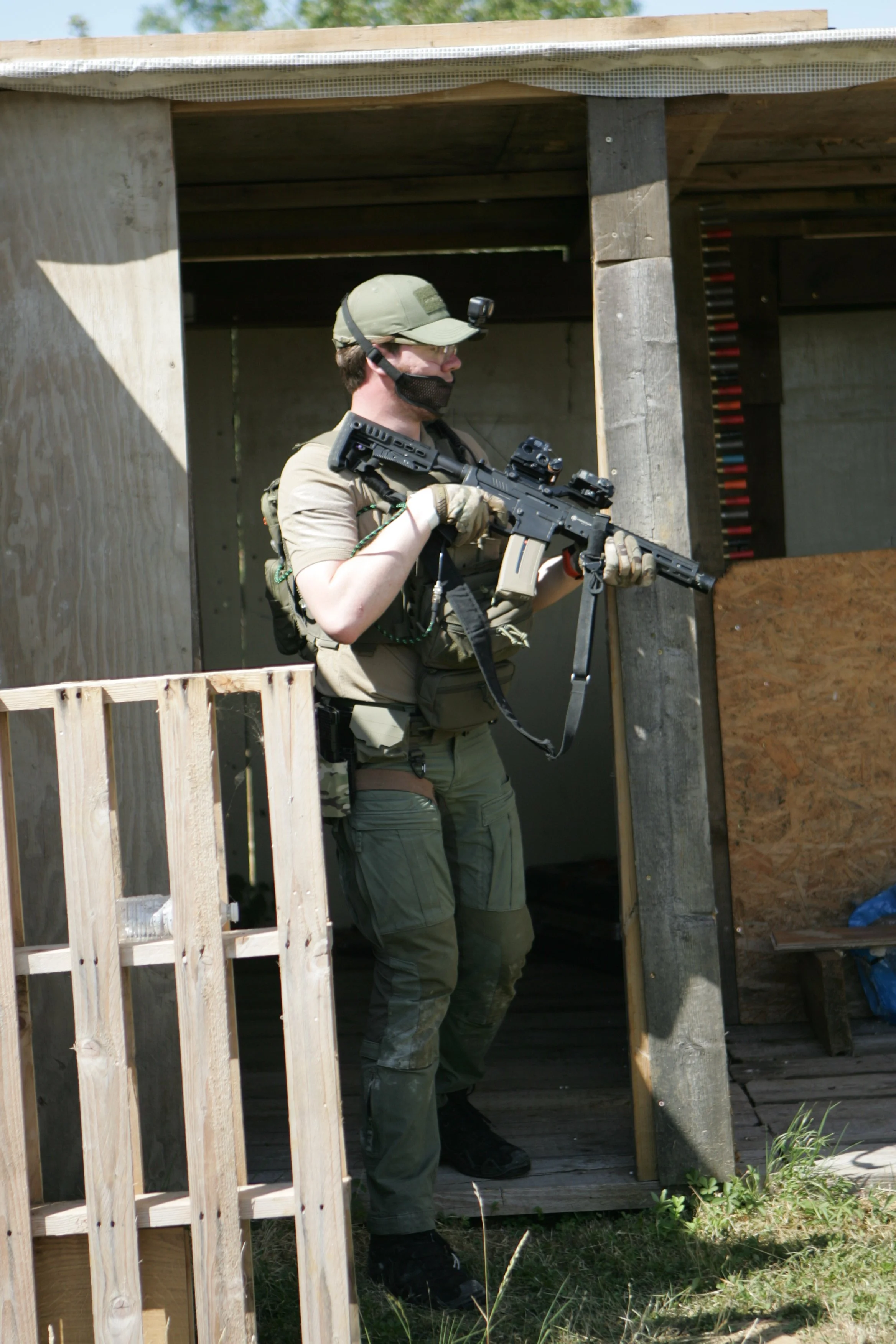 A man in tactical gear holding a firearm during a training or shooting exercise in a wooden structure outdoors.