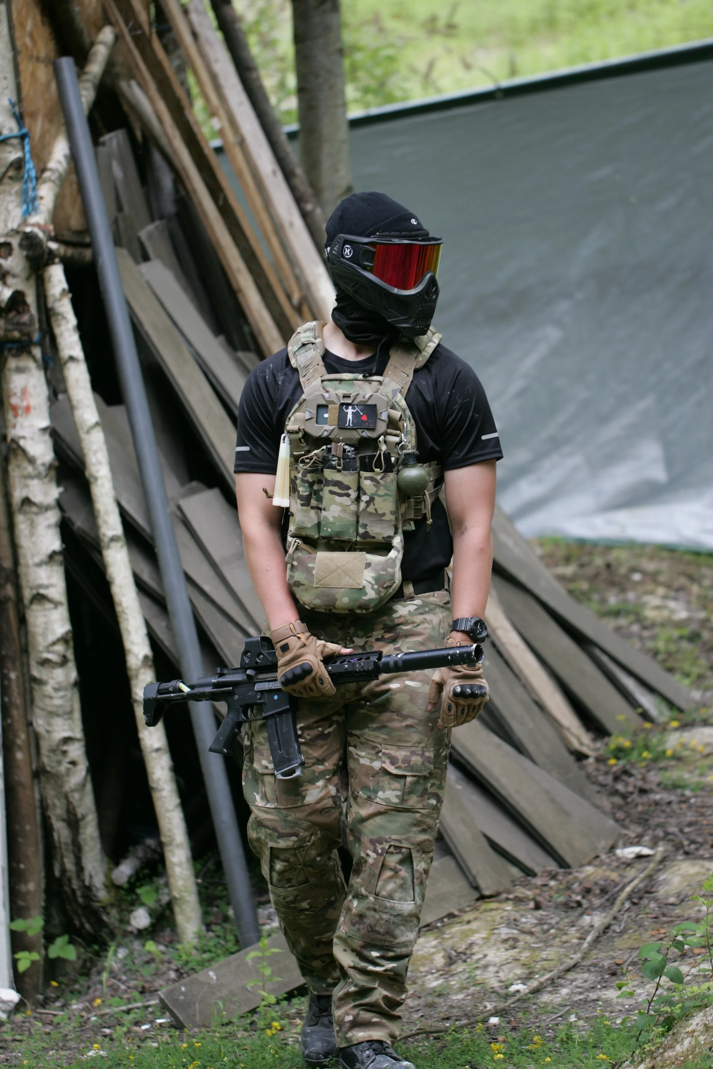 A person dressed in camouflage military or tactical attire, wearing gloves, a tactical vest, and a black face cover with a visor, holding a gun, standing outdoors near a wooden structure with logs and a plastic tarp, surrounded by trees and greenery.