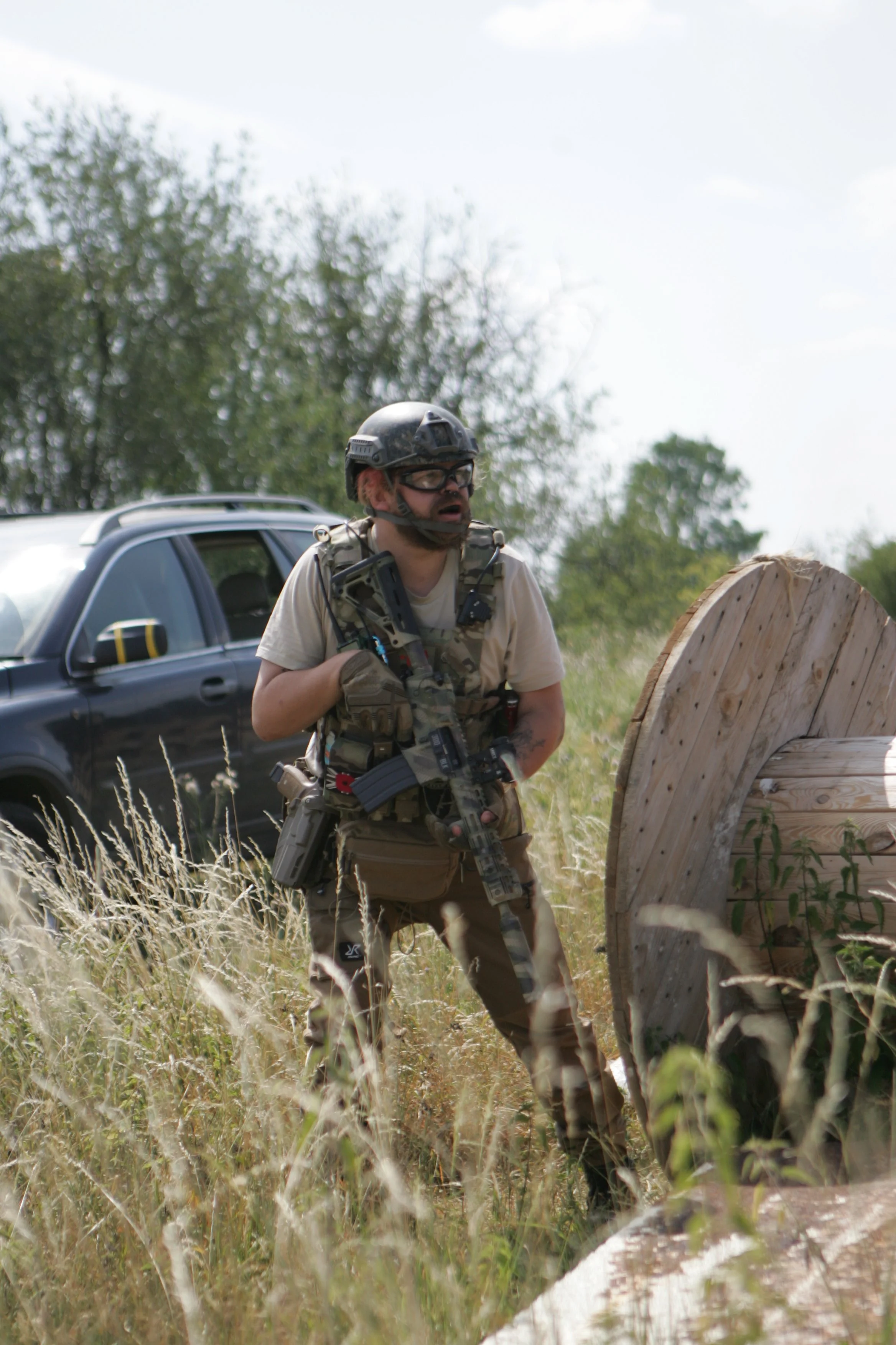 A man wearing military gear and a helmet, holding a weapon, standing outdoors near tall grass, with a car and trees in the background.