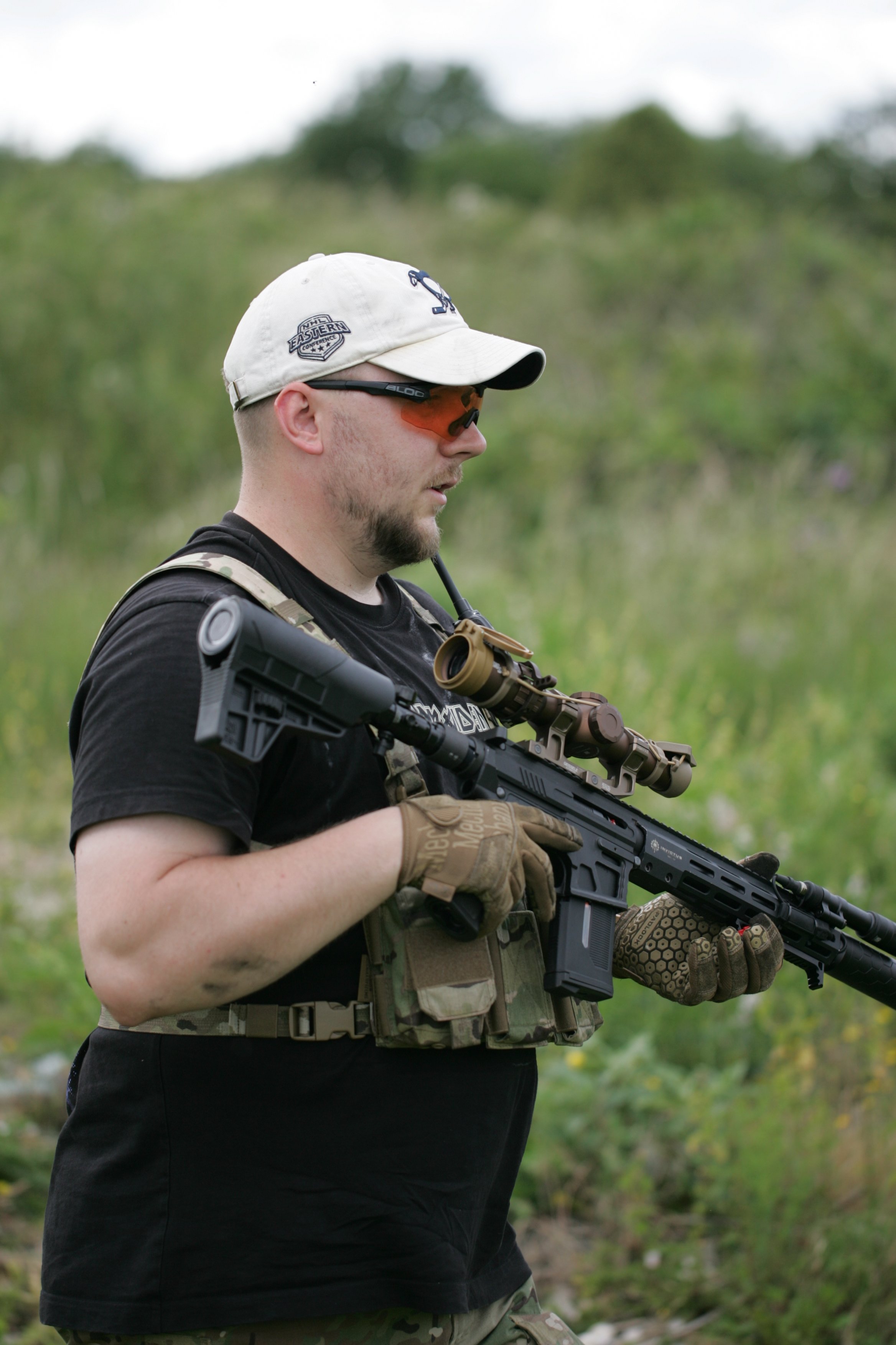 A man in a beige cap, tinted glasses, black shirt, and camouflage pants holding a rifle outdoors in a grassy area.