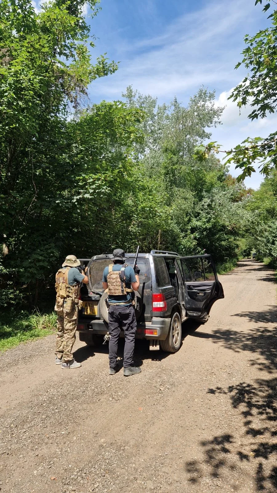 Two armed men in tactical gear standing next to an open SUV on a dirt road surrounded by dense green trees under a blue sky.