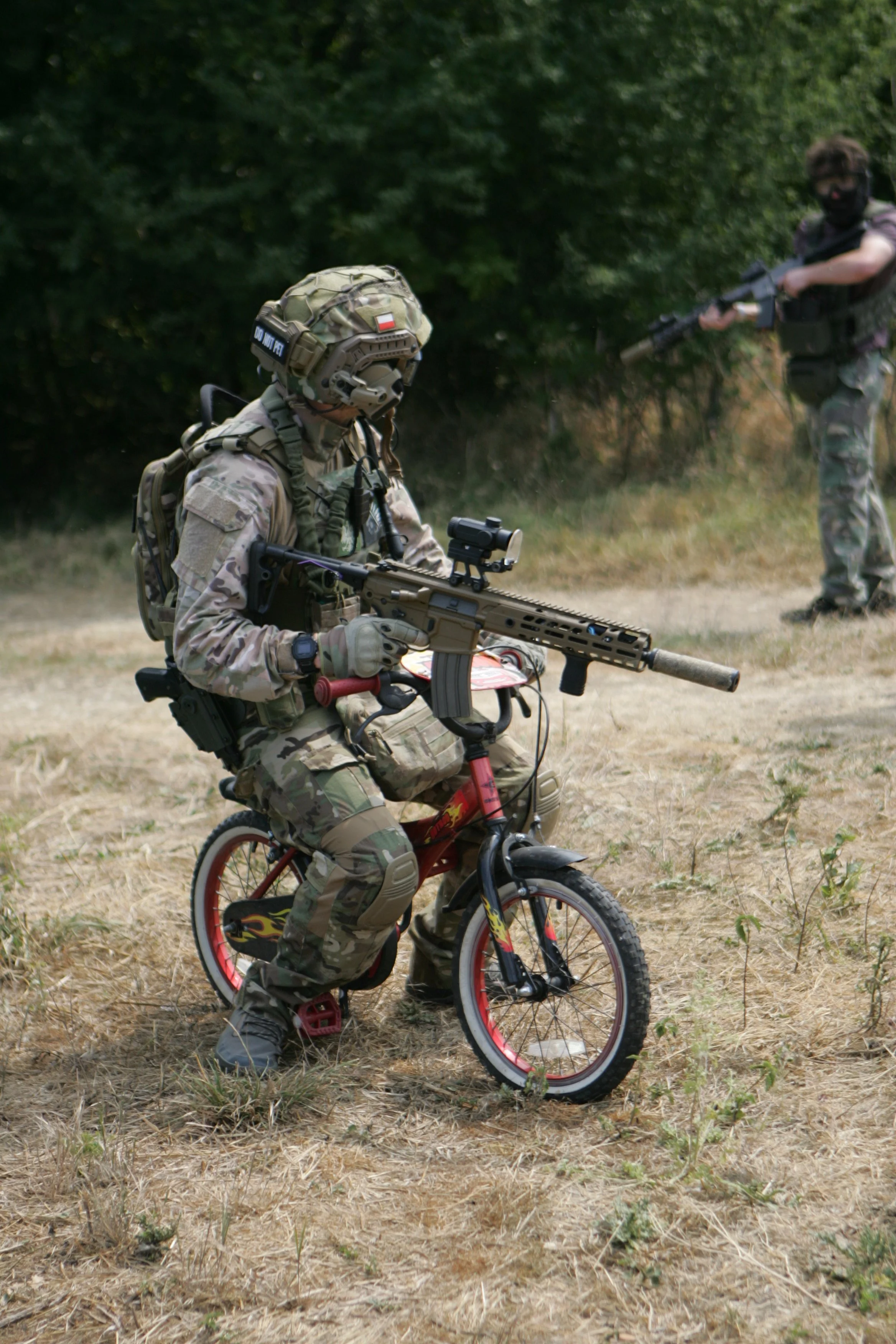 Soldiers in camouflage gear, one kneeling with a rifle and a child on a small bike, another standing with a rifle in a wooded outdoor area.