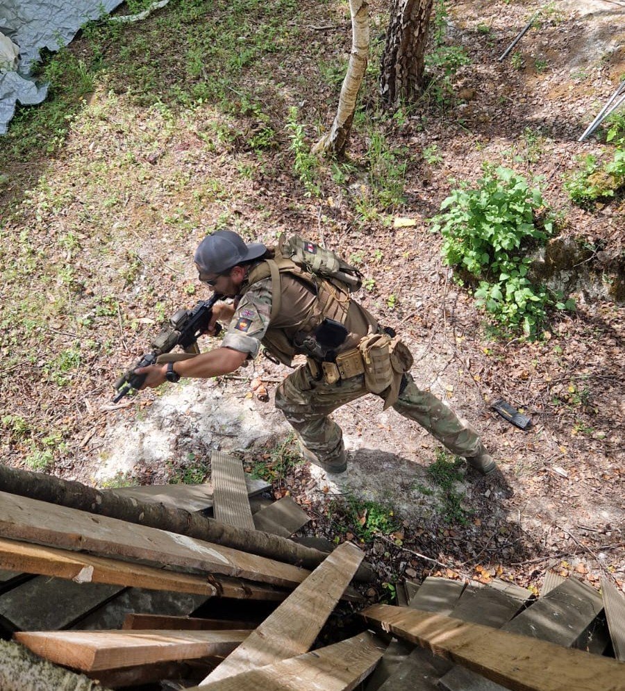 A soldier wearing camouflage gear, a helmet, and tactical equipment is aiming a rifle behind cover in a wooded outdoor area.