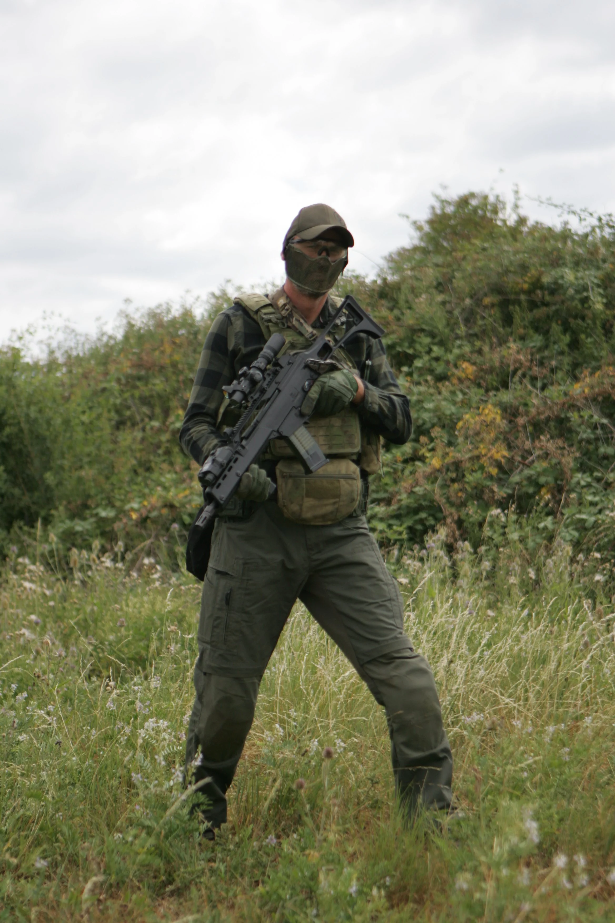 A person dressed in tactical gear, including a camouflage face mask and gloves, holding a rifle, walking through a grassy field with bushes and trees in the background.