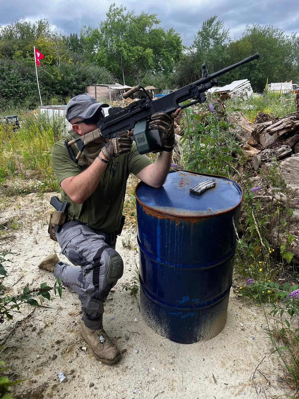 A man in tactical gear kneeling behind a blue metal barrel, aiming a large rifle with a scope outdoors. The scene features overcast skies, green trees, and some debris and plants around.