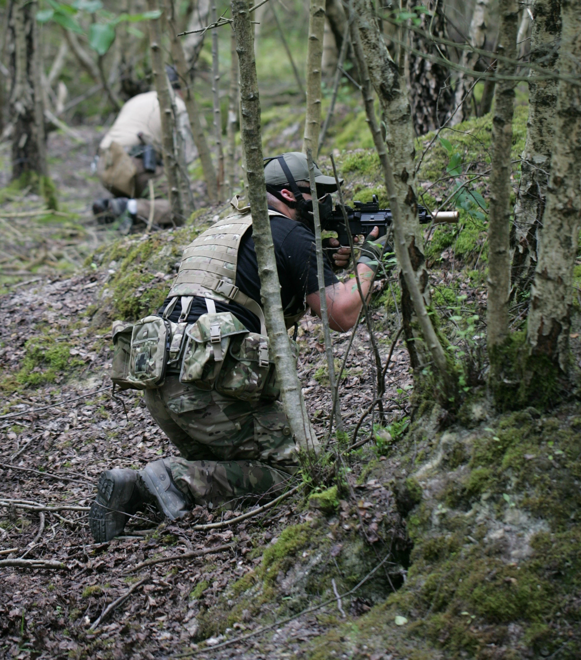 Soldier in camouflage uniform and tactical gear, kneeling behind trees and aiming a rifle in a forest.