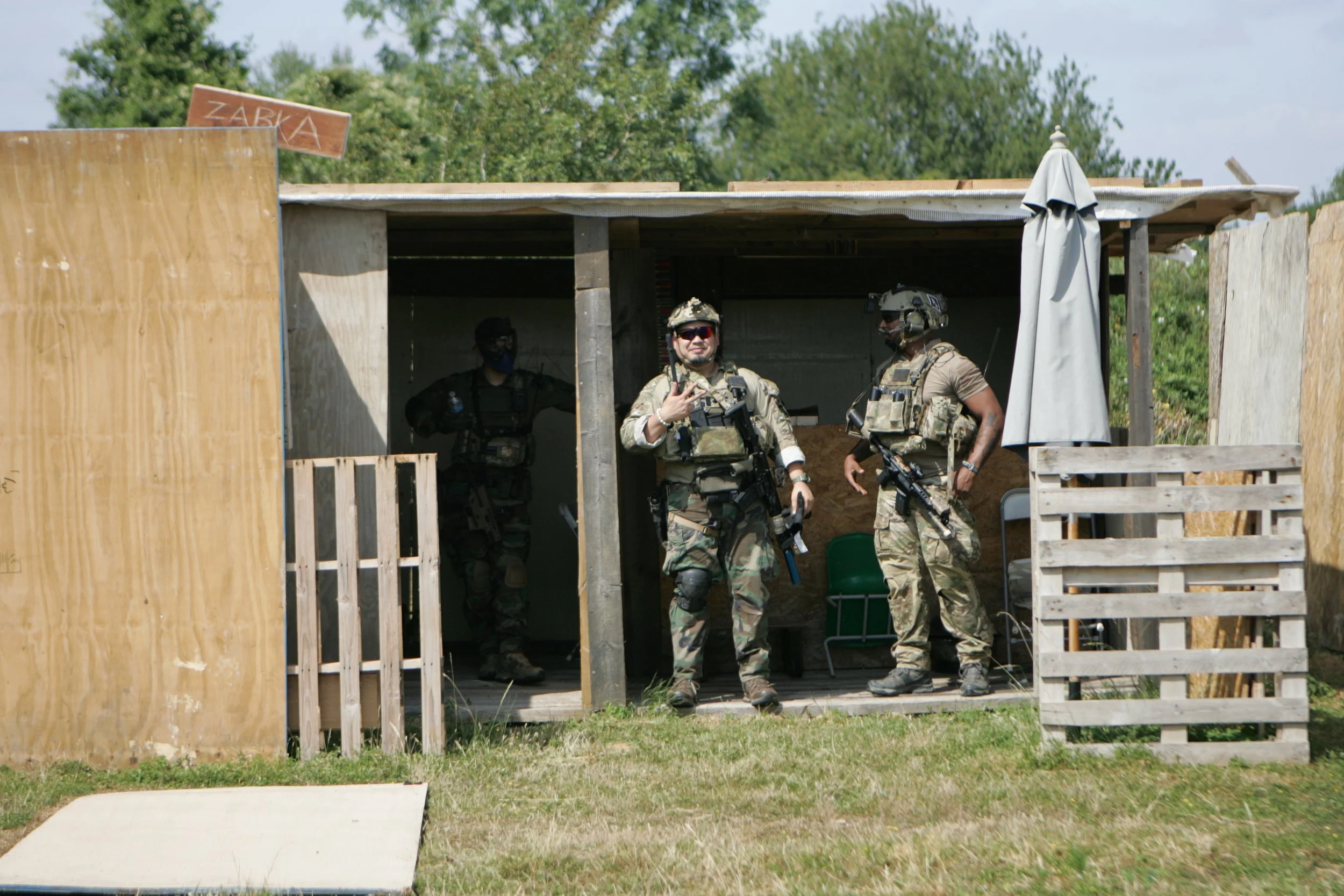 Three soldiers in camouflage uniforms and tactical gear standing outside a wooden structure with a sign that says 'ZABKA'. One soldier is making a peace sign and all are holding rifles.