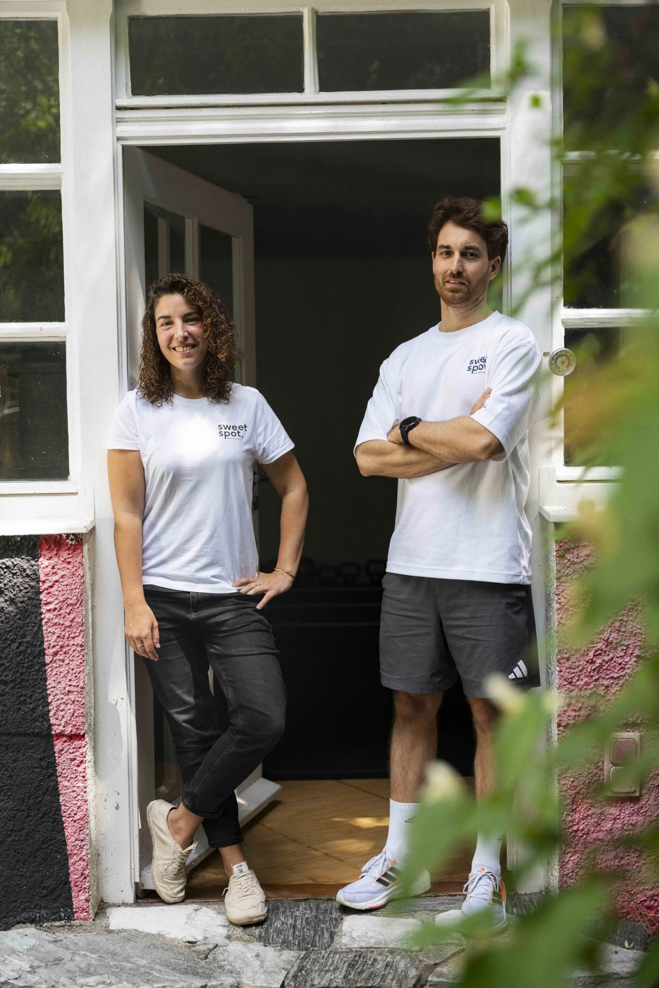 Zwei Menschen, ein Mann und eine Frau, stehen in einem Eingang und posieren für die Kamera. Beide tragen weiße T-Shirts mit dem Logo ‚sweet spot‘. Der Mann hat dunkle, lockige Haare, einen Bart und trägt Shorts. Die Frau hat lockiges braunes Haar, trägt schwarze Hosen und Sneakers. Sie lachen und scheinen sich in einem gemütlichen, häuslichen Umfeld zu befinden.