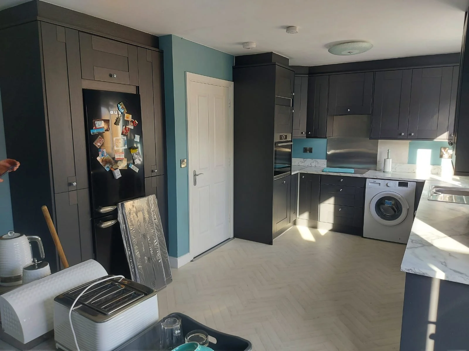 Modern kitchen with dark grey cabinets, white marble countertops, and stainless steel appliances, including a washing machine, oven, and stovetop. There is a white door, a black refrigerator with magnets, and a window letting in natural light. Items 