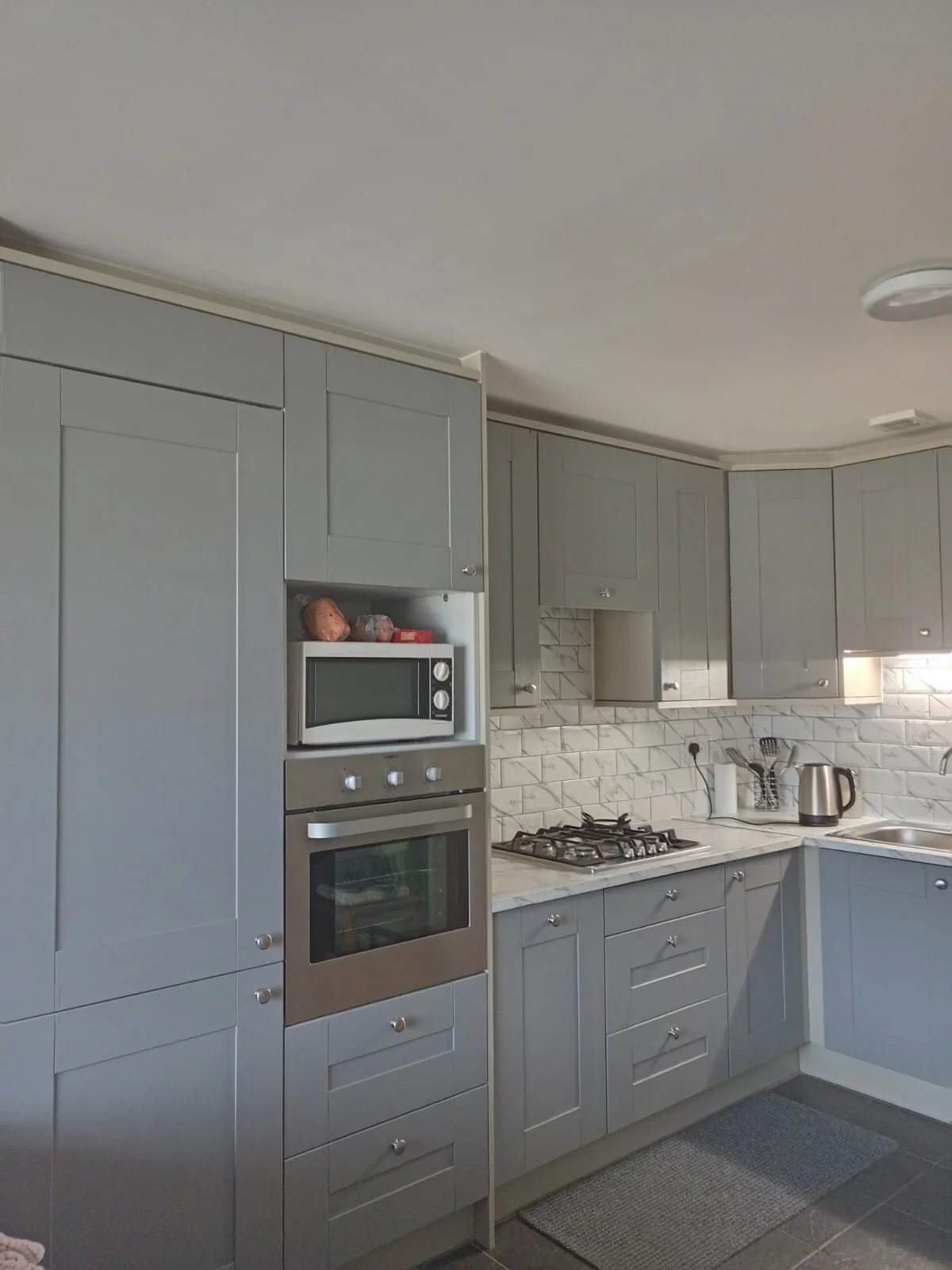 Modern kitchen with gray cabinets, marble countertops, and white subway tile backsplash; includes microwave, oven, stove, kettle, and cooking utensils.