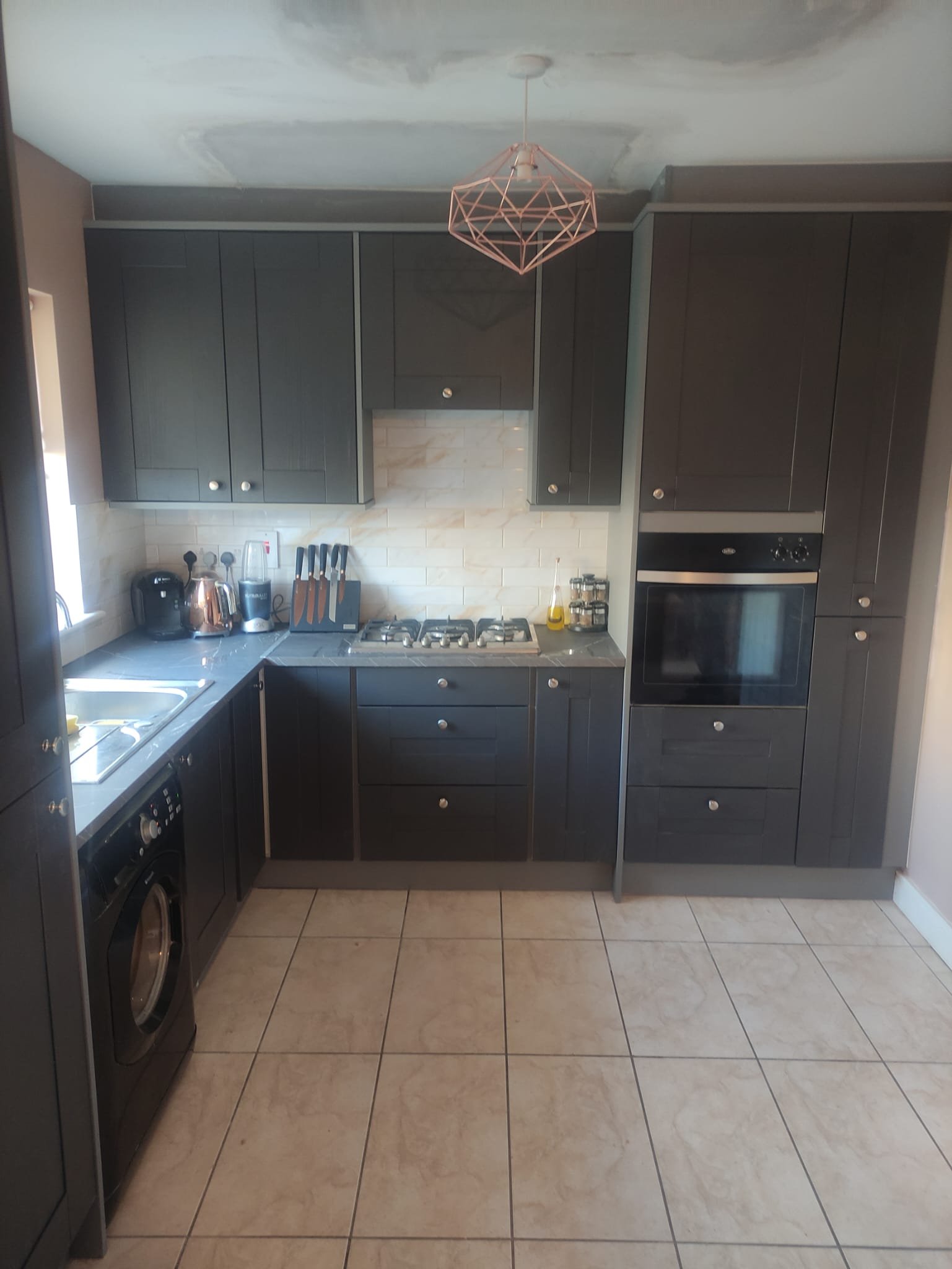 Modern kitchen with dark cabinets, built-in oven, and gas stove. Items on counter include knives, a kettle, and spices. Tiled floor and beige backsplash.