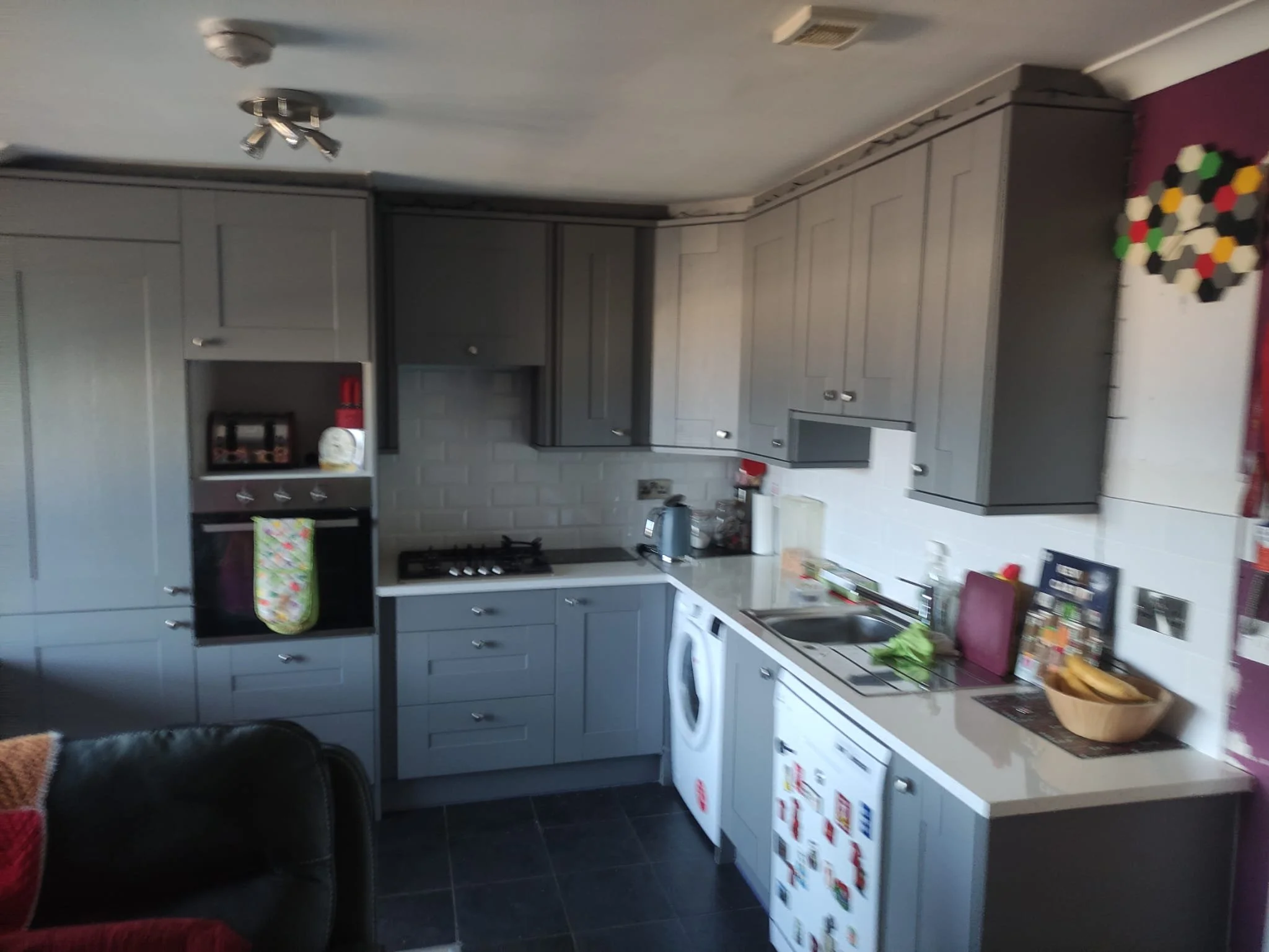 A kitchen with gray cabinets, a white countertop, and various appliances including a stove, oven, and microwave. There is a refrigerator decorated with magnets, a rice cooker, and a bowl of bananas on the counter. The floor appears to be dark tile, and the ceiling has a small fan and light fixture.