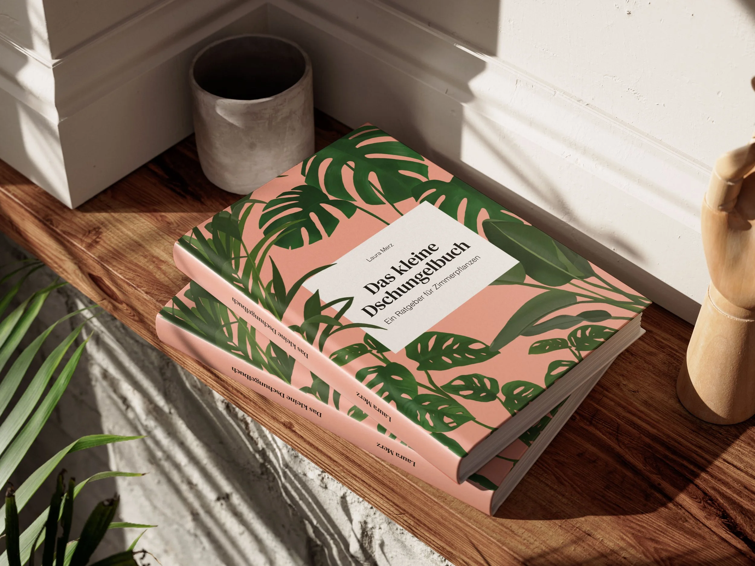 Ein aufgeschlagenes Buch mit dem Titel 'Das kleine Pflanzen-Quizbuch' liegt auf einem Holztisch neben einer Pflanze. Im Hintergrund stehen weiße Wände und eine Pflanze mit grünen Blättern.