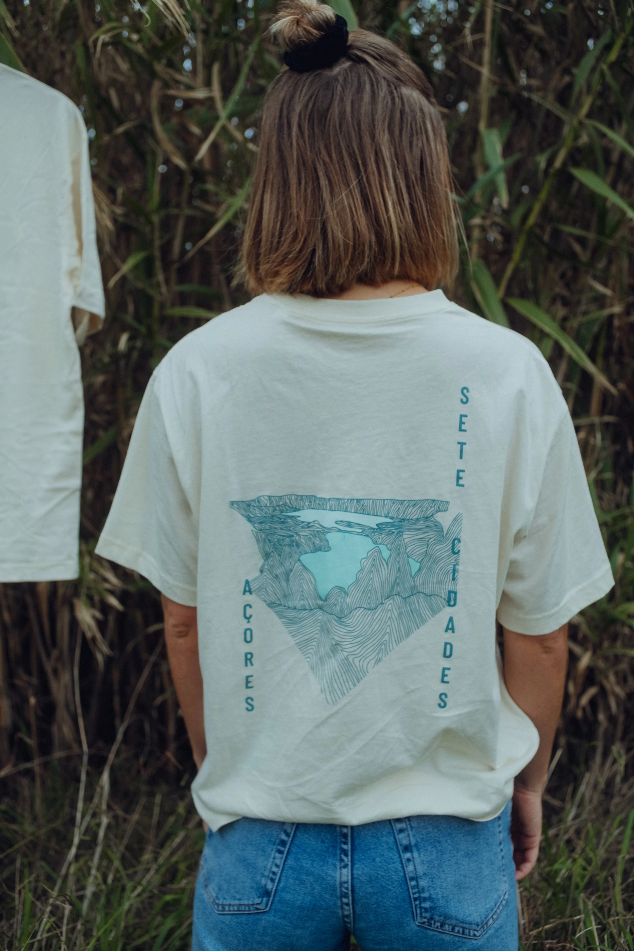 Rückansicht einer Person mit braunen, mittellangen Haaren, die eine weiße T-Shirt trägt, auf dem ein stilisiertes Landschaftsbild und die Worte 'Sete' und 'Cores' zu sehen sind, umgeben von Grün und Büschen.