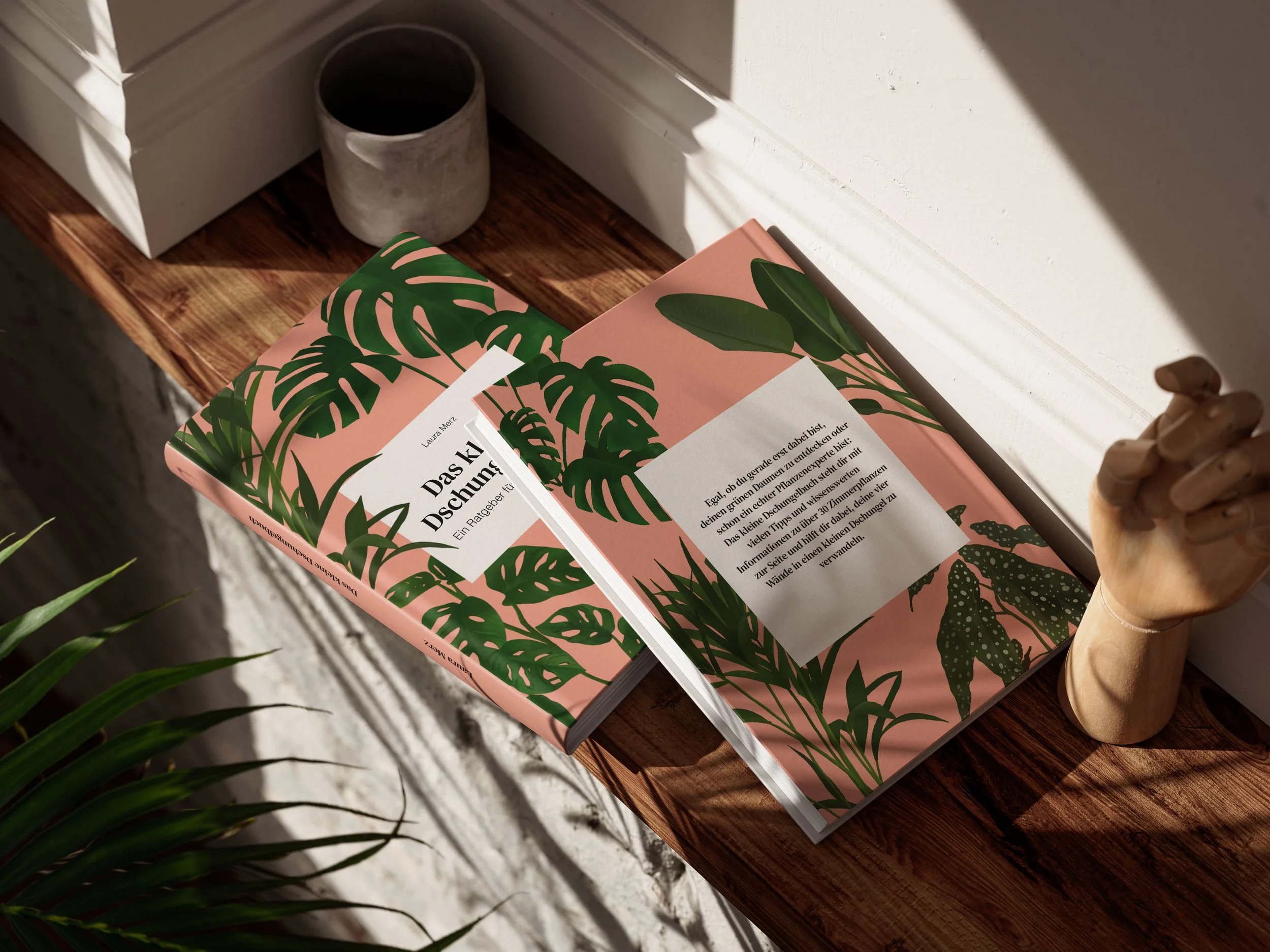 Ein pinkfarbendes Buch mit grünen Pflanzenmotiven liegt auf einem Holztisch, daneben ein grauer Blumentopf und ein kleiner Holzwürfel. Schatten fallen auf die weiße Wand im Hintergrund.