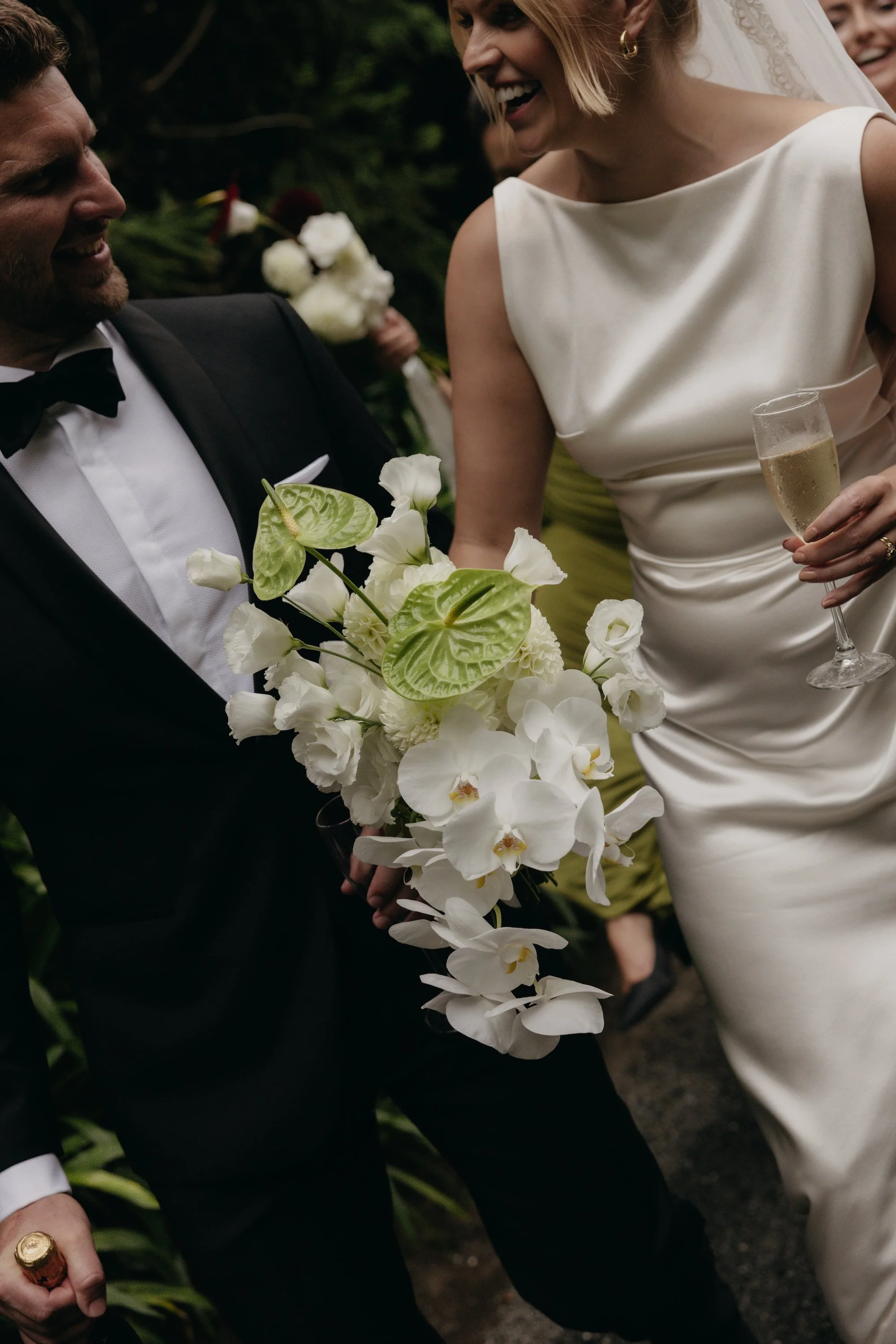 A bride and groom at their wedding, smiling and holding a wedding bouquet with white flowers and green leaves, with a bridesmaid holding a champagne flute in the background.
