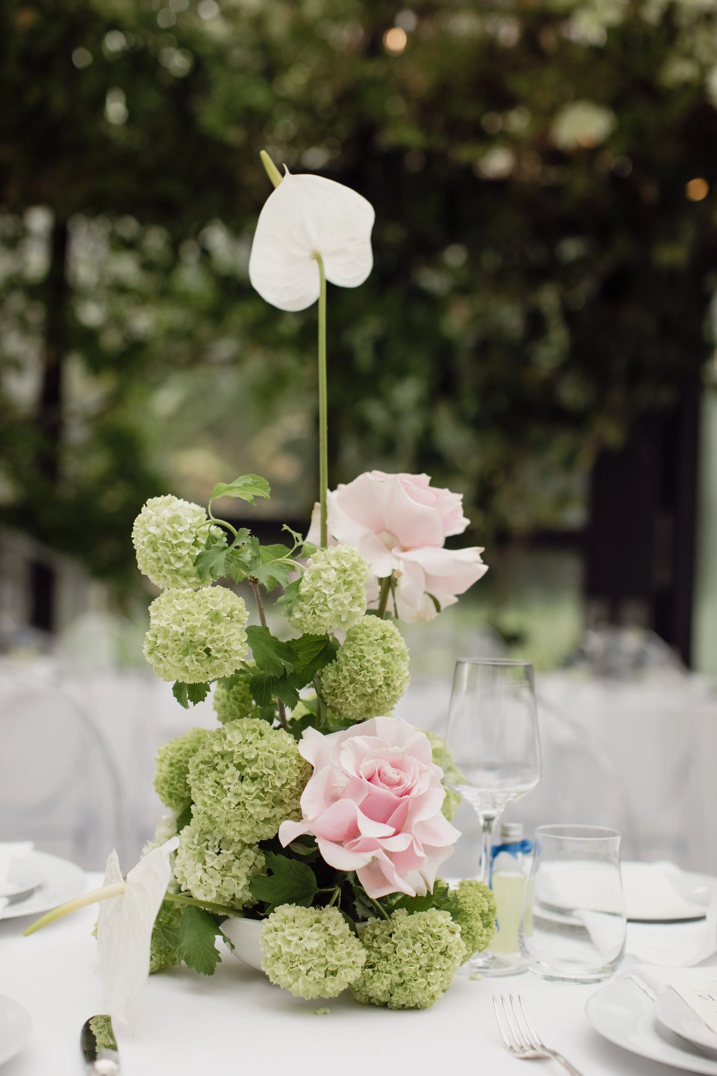 Elegant floral centerpiece featuring white anthuriums, pale pink roses, and white hydrangeas on a dining table at an outdoor event.