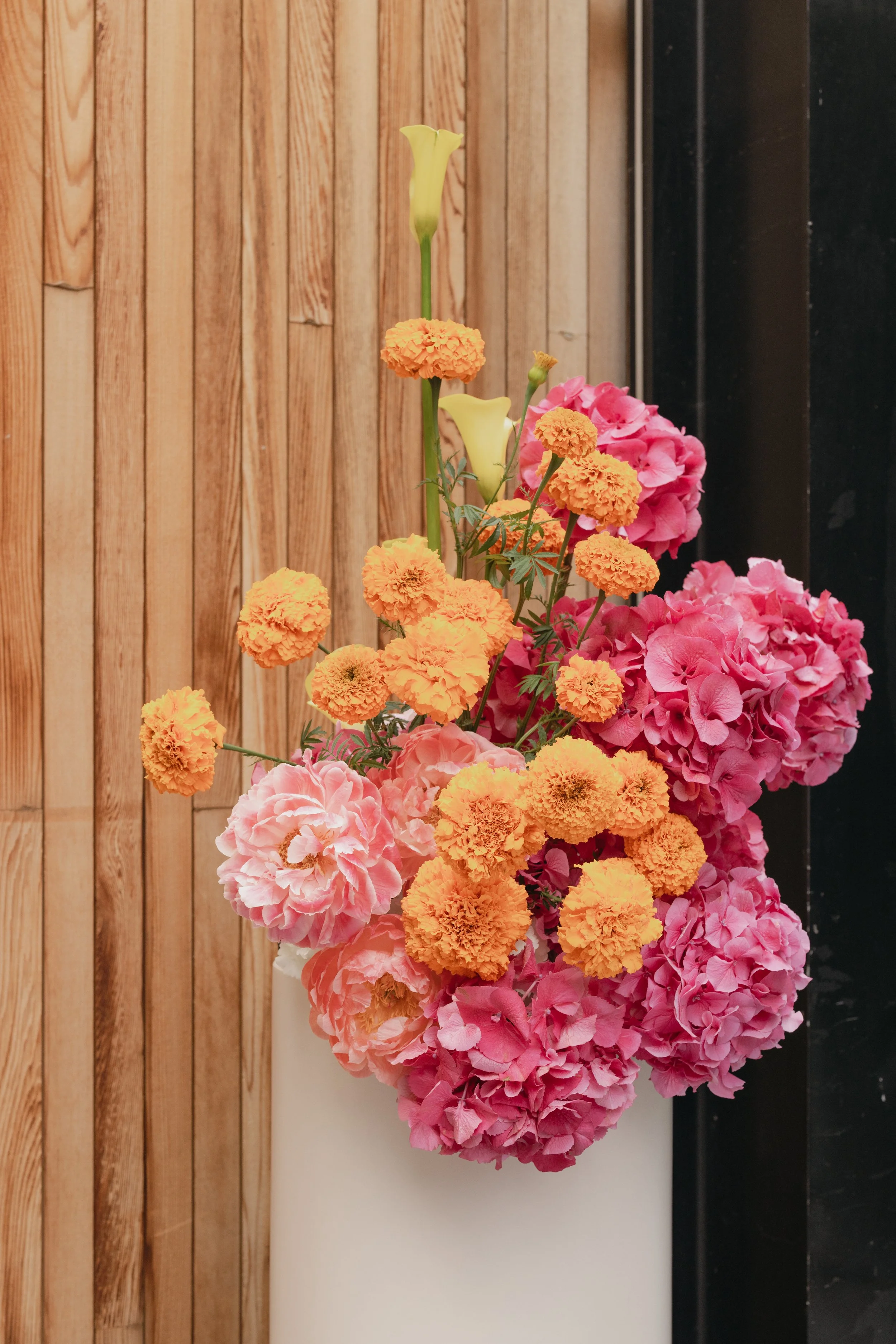 A bouquet of pink hydrangeas, orange marigolds, and yellow calla lilies against a backdrop of wooden panels and a black surface.