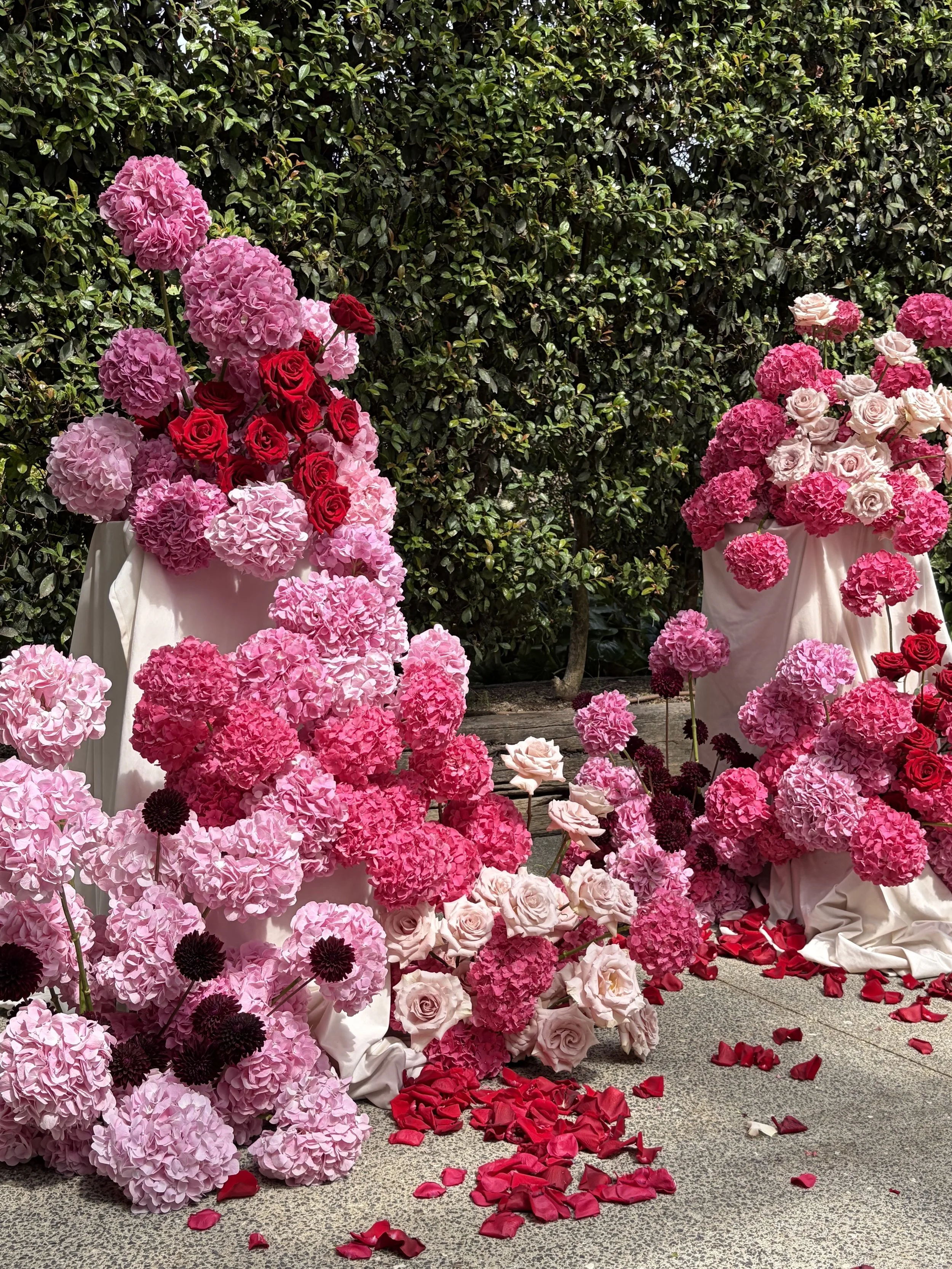 Artificial flower arrangement with pink hydrangeas, red roses, and pale pink roses in white draped vases, with scattered red petals on the ground, set against a green leafy hedge background.