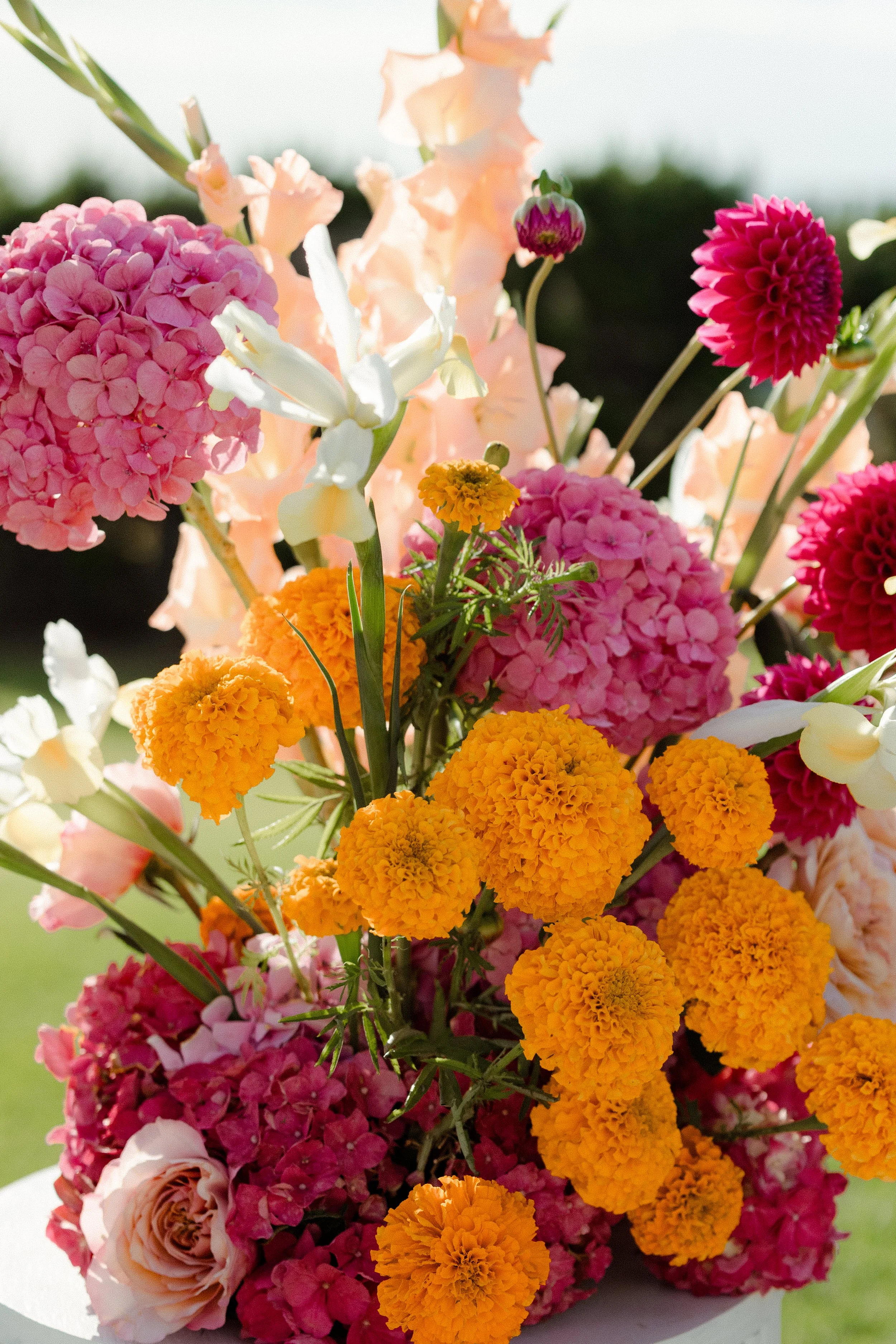 A colorful bouquet of various flowers including pink hydrangeas, orange marigolds, white orchids, pink dahlias, and pale peach roses, arranged outdoors.